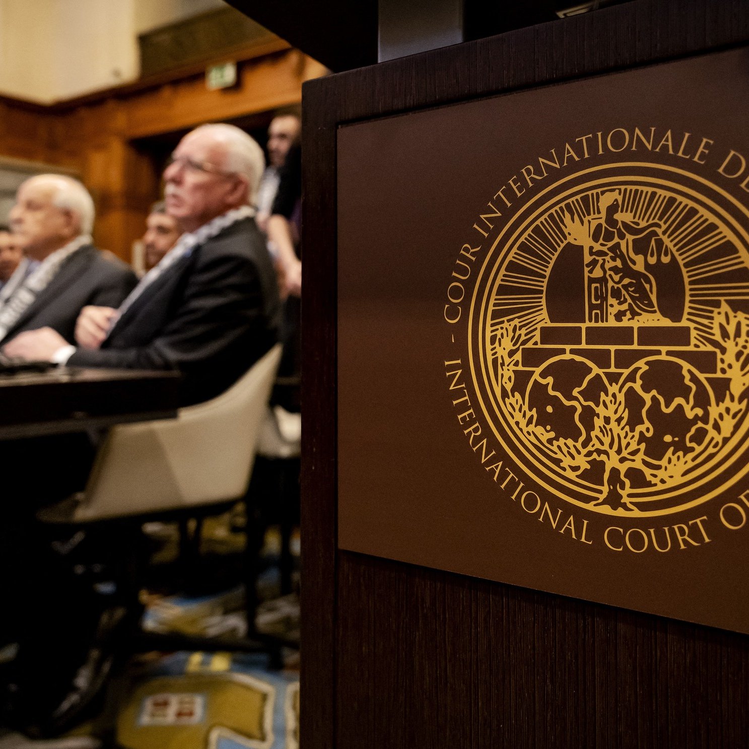 L'immagine mostra un incontro che si svolge in un ambiente formale, probabilmente in un'aula di tribunale. In primo piano si vede un simbolo dell'International Court of Justice (Corte Internazionale di Giustizia), con dettagli dorati che rappresentano l'autorità e la giustizia. Nel background si notano alcune persone sedute, molte delle quali sono visibili di spalle. L'atmosfera è seria e professionale, suggerendo un'importante deliberazione legale.
