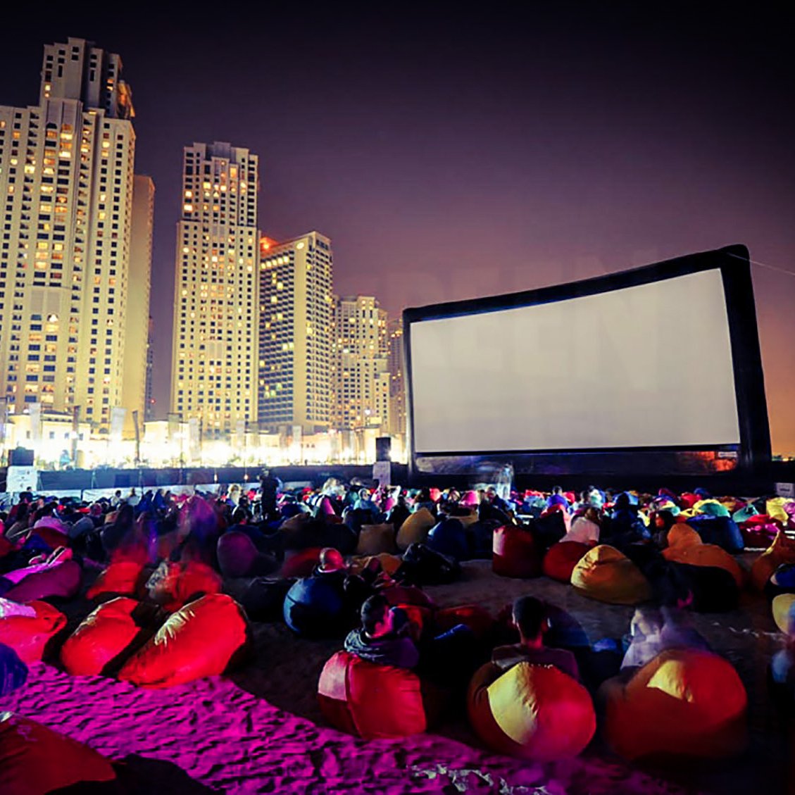 L'image montre une scène de cinéma en plein air sur une plage, animée pendant la nuit. Un grand écran est installé, et des groupes de personnes s'asseyent sur des poufs colorés, principalement de couleur rouge. En arrière-plan, on peut voir des gratte-ciels illuminés, créant une atmosphère vibrante et moderne. L'éclairage doux et les ombres ajoutent à l'ambiance festive du lieu.