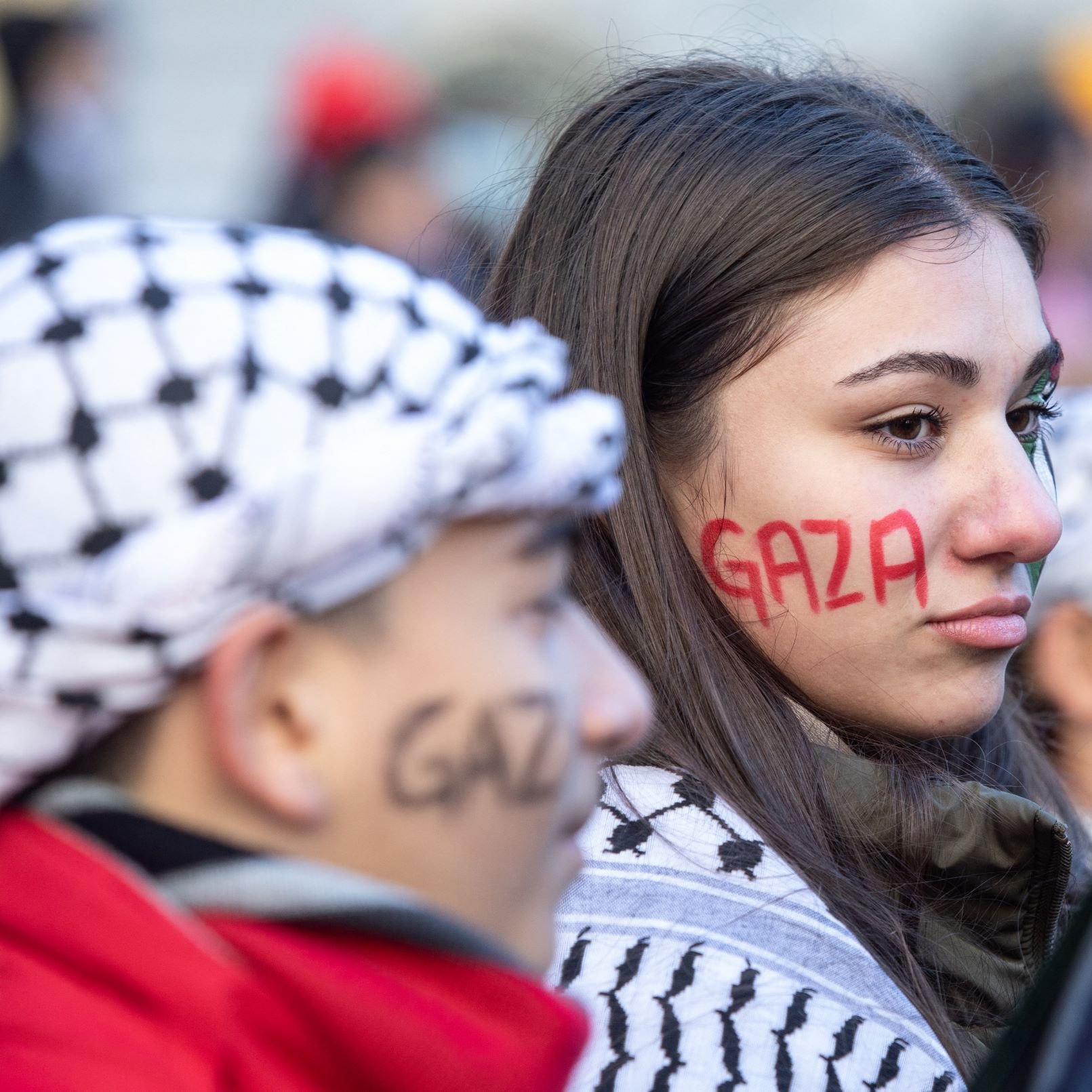 در تصویر، چند نفر در حال شرکت در یک تجمع یا اعتراض به چشم می‌خورند. یک دختر جوان با چهره‌ای جدی و نگران، روی صورتش نوشته‌ای به رنگ قرمز دارد که "Gaza" به نمایش گذاشته شده است. او همچنین شال یا لباسی با طرح‌های خاص به تن دارد. در کنار او، پسرکی نیز دیده می‌شود که روی صورتش نوشته‌ای مشابه به رنگ سیاه دارد. فضای کلی تصویر نشان‌دهنده شور و نشاط در حمایت از مسئله‌ای است که به نظر می‌رسد به غزه مربوط می‌شود. دیگر افراد نیز در پس‌زمینه حضور دارند و فضا به وضوح یک تجمع اجتماعی را تداعی می‌کند.