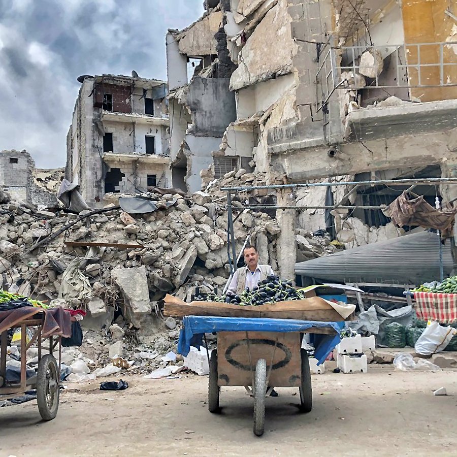 L'image montre une scène d'un marché ou d'une vente de légumes dans un environnement dévasté, avec des bâtiments en ruine en arrière-plan. On voit deux personnes, probablement des vendeurs, avec des charrettes remplies de légumes, comme des courgettes ou des poivrons. L'atmosphère est chargée, avec des nuages obscurcis qui semblent se rassembler, ajoutant à la gravité de la situation. Les décombres et les bâtiments effondrés indiquent une zone touchée par un conflit ou une catastrophe.