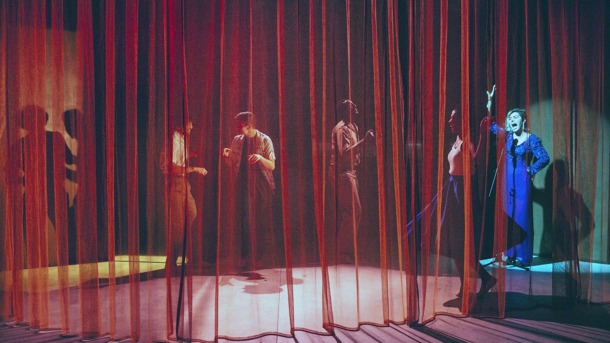 L'image montre une scène de théâtre avec des rideaux en velours rouge et orange qui créent une atmosphère intimiste. On aperçoit plusieurs silhouettes de personnes derrière les rideaux, engagées dans des activités qui semblent dynamiques et expressives. L'un des personnages, visible à droite, porte un costume bleu et semble gesticuler avec enthousiasme. Les jeux de lumière entre les rideaux ajoutent une ambiance dramatique à la scène, accentuant les mouvements des acteurs.