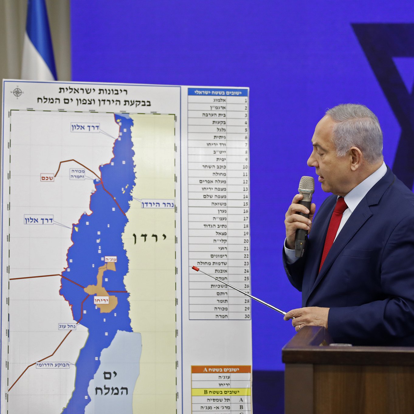 در این تصویر، یک مرد در حال ارائه یک نقشه است. او با یک میلیا بیل کوچک در دست، به نقطه‌ای روی نقشه اشاره می‌کند. نقشه در پس‌زمینه شامل خطوط و علامات جغرافیایی است. پشت او یک علامت بزرگ ستاره داوود دیده می‌شود و فضای سالن به‌نظر می‌رسد رسمی و مرتبط با یک رویداد یا کنفرانس باشد. لباس مرد رسمی به نظر می‌رسد و او به‌وضوح در حال بیان نکات کلیدی است.