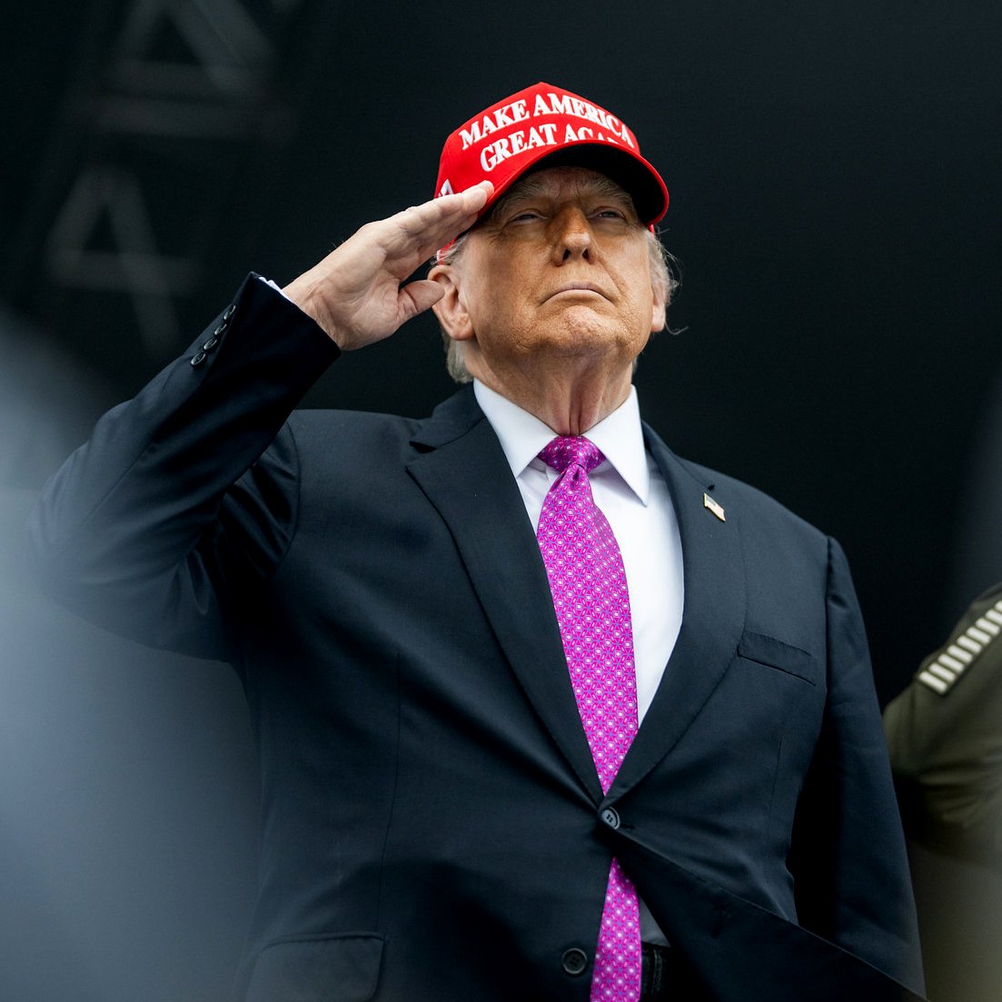 Uomo in abito scuro e cravatta viola, con berretto rosso, fa saluto militare.