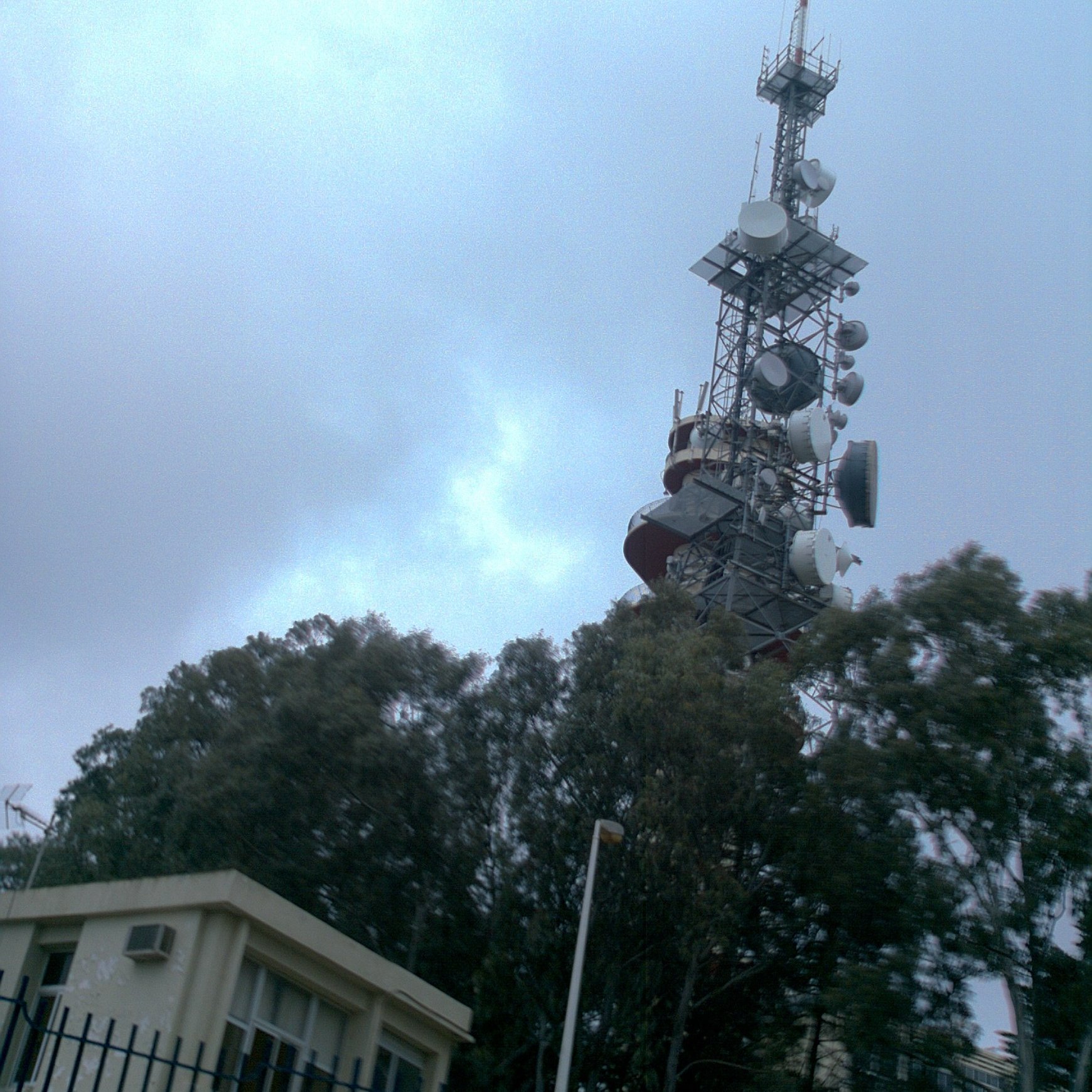 L'image montre une tour de télécommunication élevée, dotée de plusieurs antennes et équipements en haut. Elle est entourée de grands arbres verts et se dresse à côté d'un bâtiment plus petit, probablement un centre d'opérations ou de maintenance. Le ciel est nuageux, ce qui donne une ambiance légèrement grise et sombre à la scène.