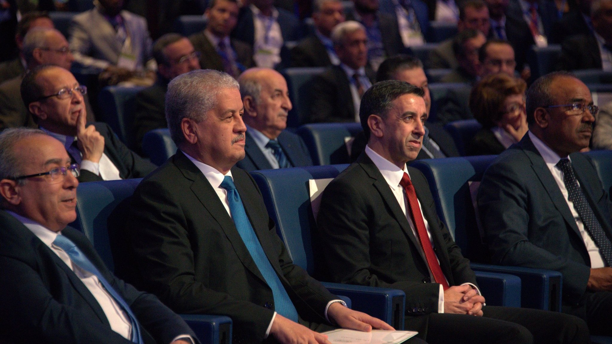 L'image montre un groupe d'hommes assis dans une salle de conférence, apparemment lors d'un événement officiel. Ils portent des costumes sombres, avec une attention particulière sur deux hommes au premier plan, l'un en costume noir avec une cravate bleue, et l'autre en costume noir avec une cravate rouge. En arrière-plan, on peut voir d'autres personnes, probablement des participants ou des invités, attentifs à ce qui se passe sur scène. L'atmosphère semble formelle et sérieuse.