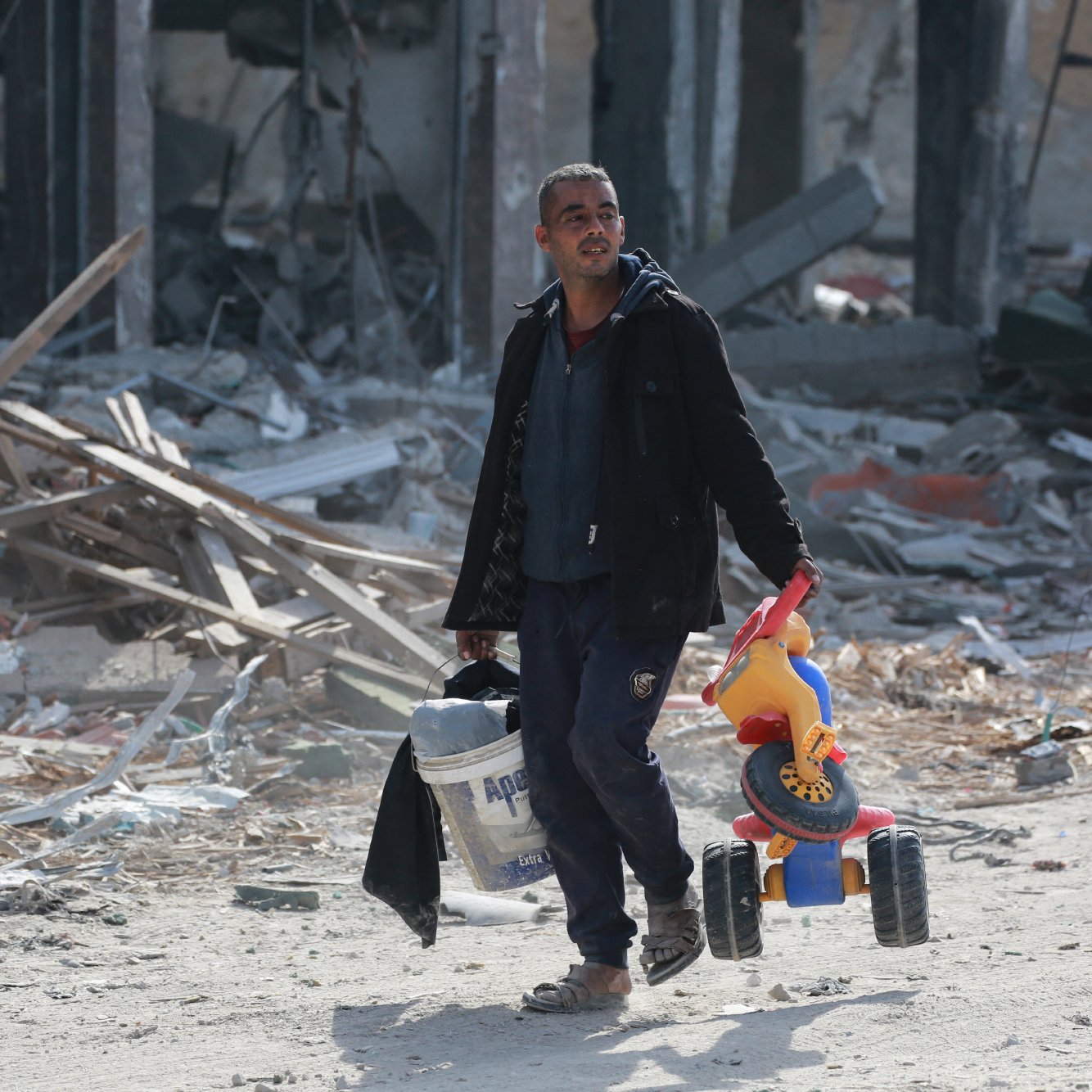 L'immagine ritrae un uomo che cammina in un'area distrutta, circondato da macerie e detriti. Indossa un abbigliamento scuro e porta con sé un secchio e un giocattolo per bambini, un veicolo colorato. L'atmosfera è di desolazione, evidenziata dai resti di edifici in secondo piano. L'uomo appare concentrato mentre avanza nella scena.