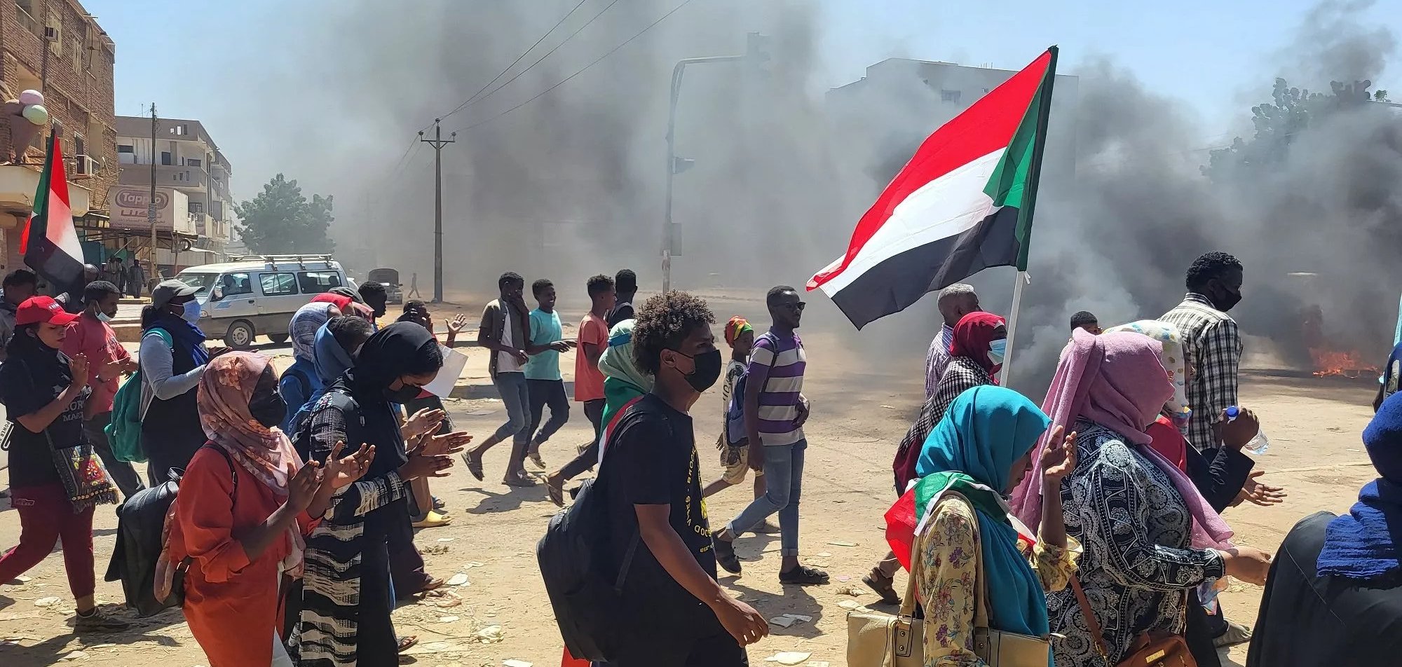 Manifestanti con bandiera del Sudan, in mezzo a fumi di incendi e tensione.