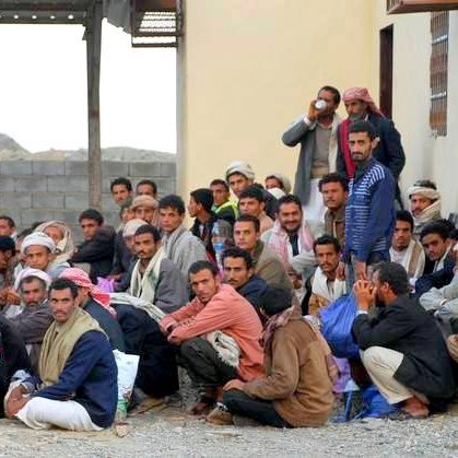 L'image montre un groupe de nombreux hommes assis au sol, semblant se regrouper près d'un bâtiment. Certains portent des vêtements traditionnels, tandis que d'autres ont des vêtements plus modernes. Ils semblent discuter ou écouter attentivement quelque chose. L'arrière-plan présente des murs en pierre et une construction simple. L'atmosphère semble calme, mais on peut sentir une certaine intensité dans leur rassemblement.