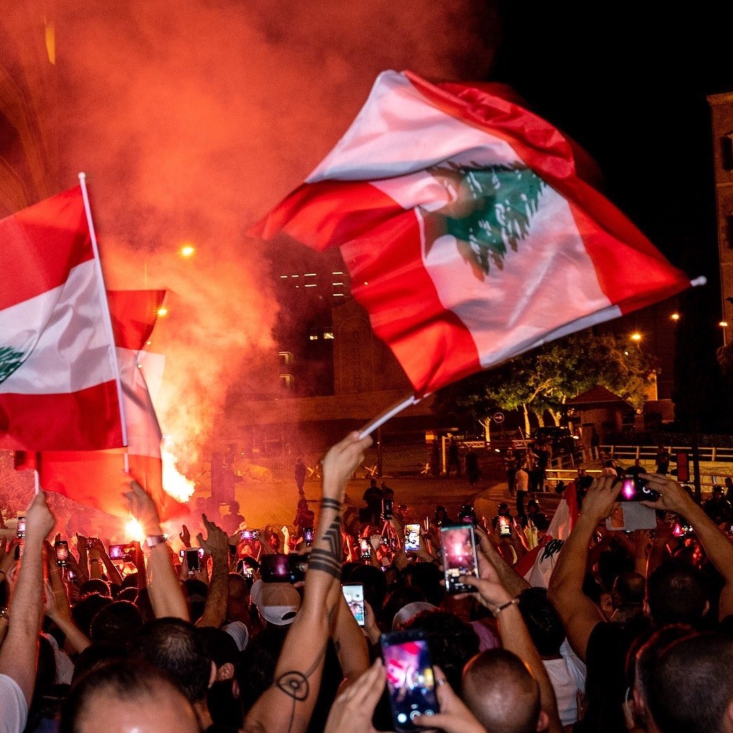 در تصویر، جمعیتی از مردم در یک تظاهرات شبانه دیده می‌شوند. افراد با پرچم‌های لبنان در دست، در حال شادی و ابراز احساسات هستند. یک شعله آتش بزرگ در پس‌زمینه به روشنایی فضای شب افزوده و به جو فعالیت و هیجان دامن می‌زند. تعداد زیادی از افراد گوشی‌های هوشمند خود را برای ثبت لحظات بالا برده‌اند. این صحنه نمایانگر شور و شوق اجتماعی و همبستگی مردم است.