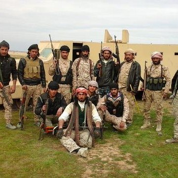 L'image montre un groupe de personnes en tenue militaire, armées et posant devant un véhicule. Ils semblent être sur un terrain en extérieur, peut-être dans un environnement désertique ou rural. Les hommes affichent diverses expressions et postures, certains sont debout tandis que d'autres sont assis. Leurs vêtements et accessoires suggèrent qu'ils font partie d'une unité militaire ou paramilitaire. En arrière-plan, on aperçoit un bâtiment et une tour d'eau.