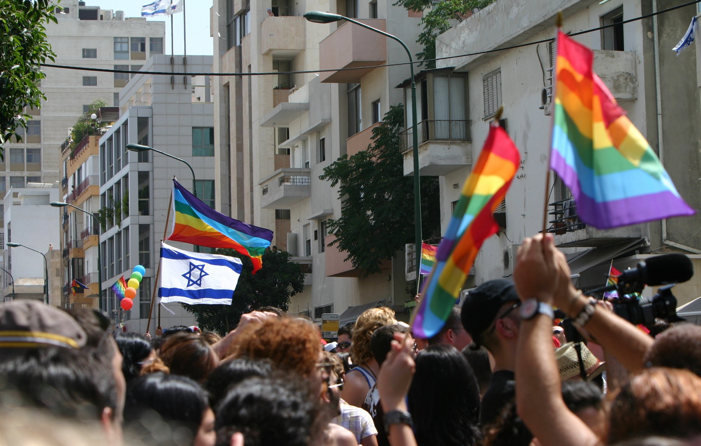 L'image montre une scène vibrante de célébration lors d'un défilé de fierté. On y voit une foule de personnes brandissant des drapeaux arc-en-ciel, symboles de la communauté LGBTQ+. Parmi eux, une grande bannière représentant le drapeau d'Israël flotte également. En arrière-plan, on distingue des bâtiments urbains, et plusieurs personnes semblent s'amuser et célébrer l'événement, créant une ambiance festive et joyeuse.
