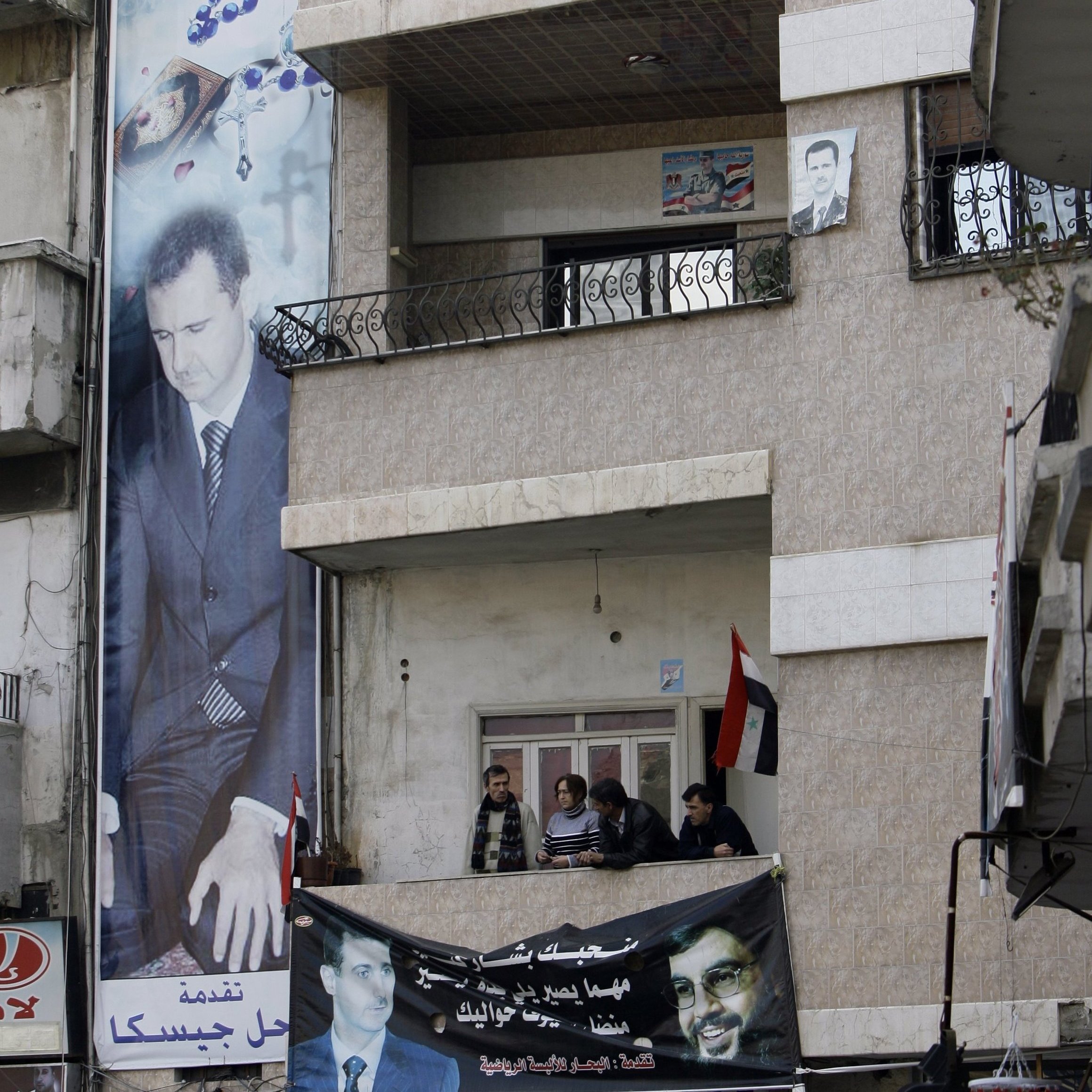 L'image montre un bâtiment avec une grande affiche représentant un homme en costume, probablement une figure politique. On peut voir plusieurs personnes sur un balcon au premier étage, observant l'extérieur. En dessous, il y a une bannière affichant un message, et des drapeaux sont visibles, ce qui suggère un contexte politique ou de soutien à une cause particulière. Les murs du bâtiment sont décorés de manière simple, et l'ambiance semble être formelle.