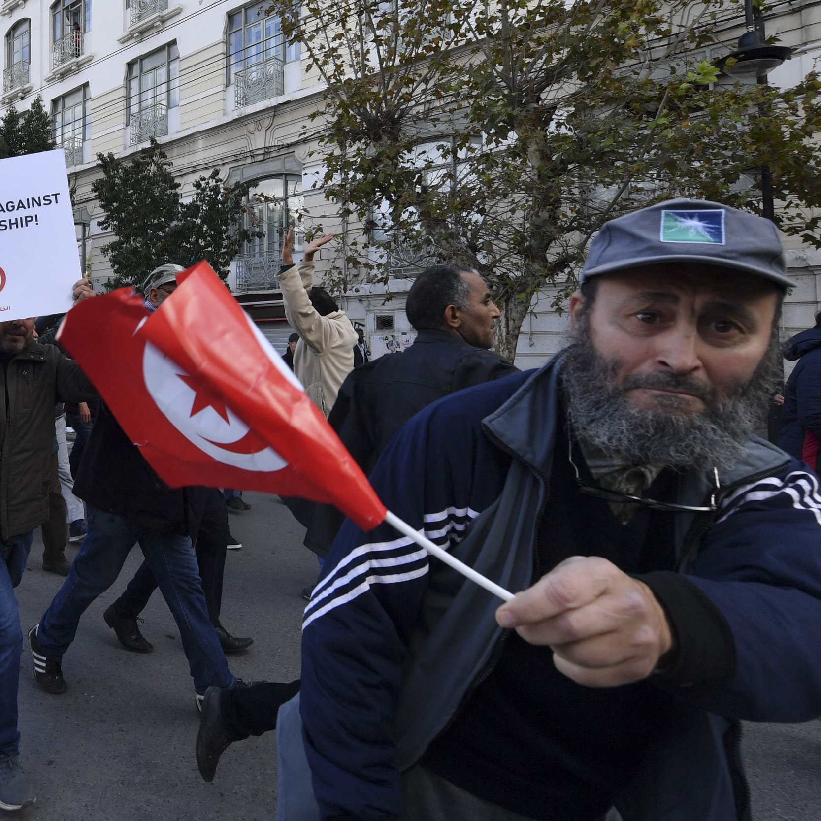 L'image montre une scène de manifestation avec des gens défilant dans la rue. Plusieurs manifestants portent des drapeaux tunisiens et tiennent des panneaux, dont l'un exprime un message contre la dictature. L'atmosphère semble énergique et engagée, avec des participants qui expriment leurs opinions et cherchent à faire entendre leur voix. Les bâtiments en arrière-plan reflètent un contexte urbain.
