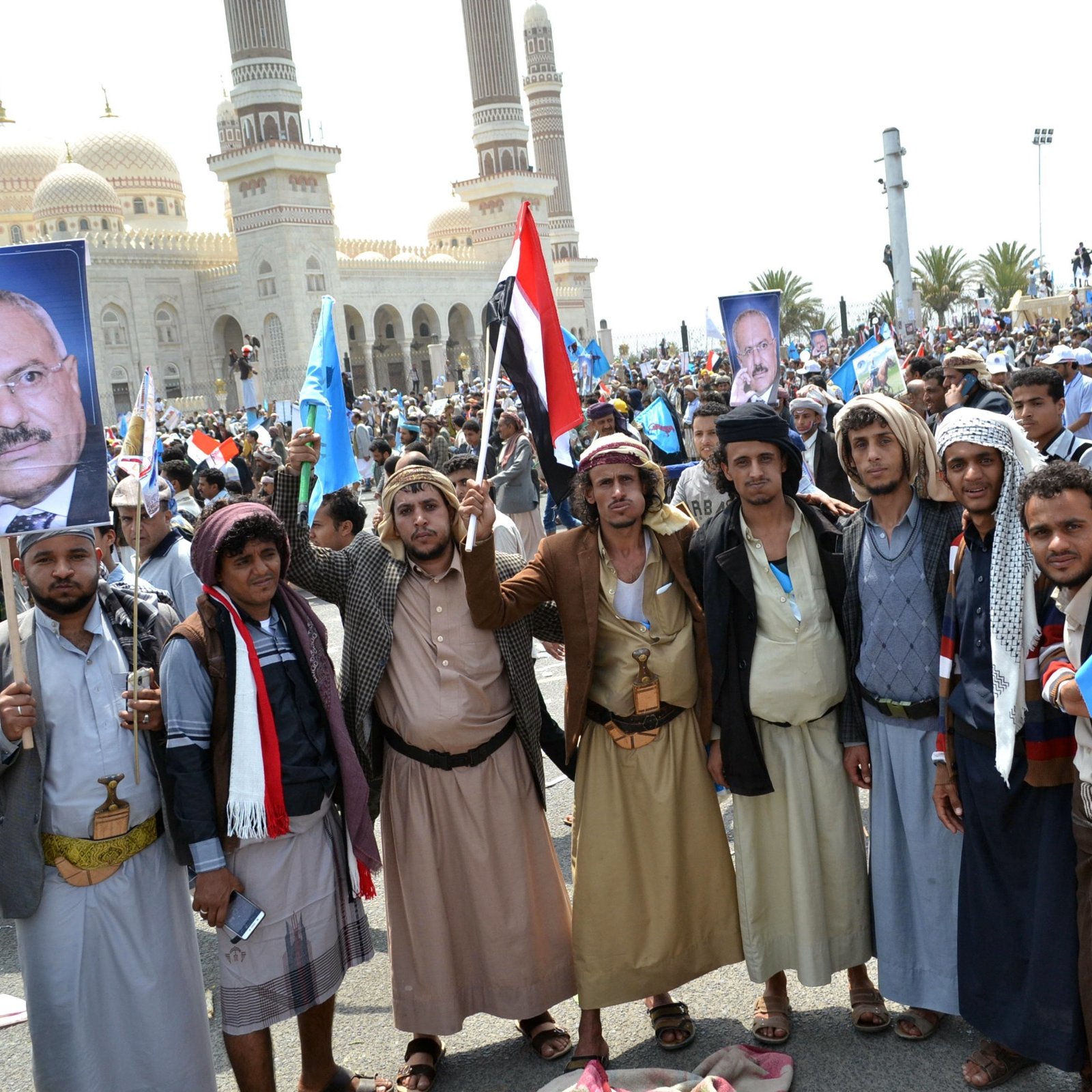L'image montre un groupe de personnes rassemblées lors d'une manifestation. Elles portent des vêtements traditionnels et tiennent des drapeaux ainsi que des pancartes, y compris une qui affiche un portrait. En arrière-plan, on peut voir une grande mosquée ou un bâtiment avec des dômes, suggérant que l'événement a lieu dans un environnement urbain. L'atmosphère semble festive et engagée, avec une forte mobilisation des participants.