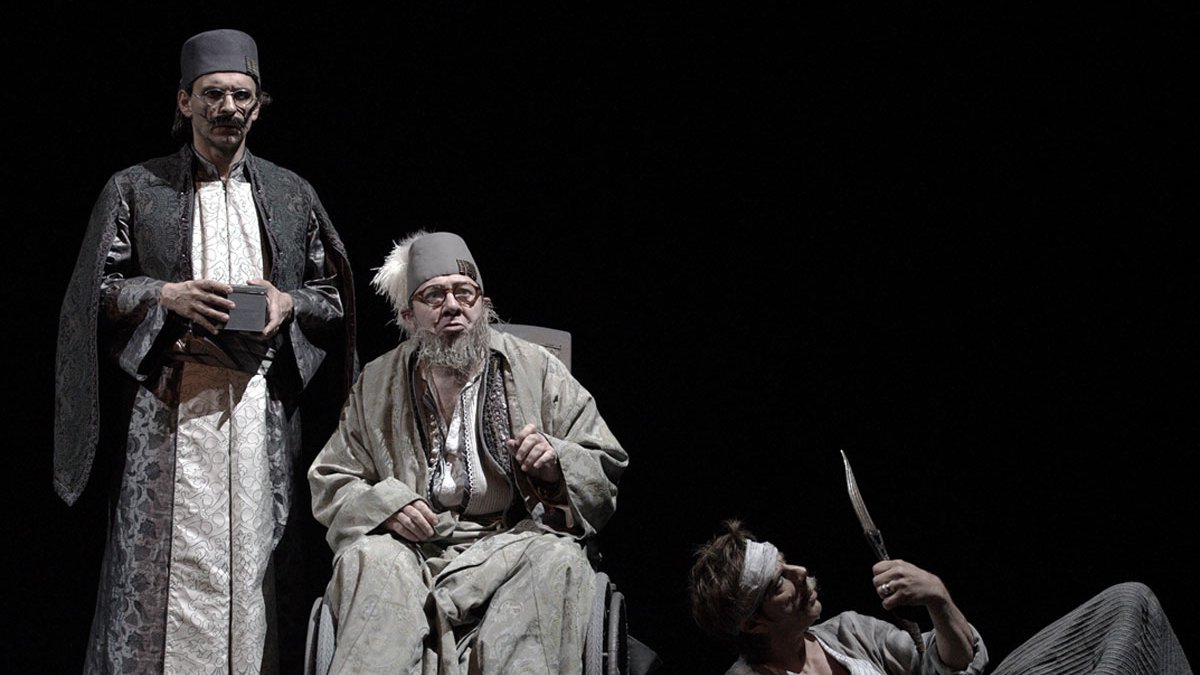 L'image représente une scène de théâtre mettant en avant trois personnages. Deux d'entre eux, habillés de façon traditionnelle et avec des vêtements sombres et ornés, semblent être en position d'autorité ou d'observation. Le troisième personnage est assis dans un fauteuil roulant, avec des vêtements plus clairs et un air pensif ou mélancolique. Il a une longue barbe grise et des lunettes. À côté de lui, un autre personnage, qui paraît être en détresse ou blessé, est au sol, tenant un objet pointu. L'éclairage est sombre, créant une atmosphère dramatique et intense.
