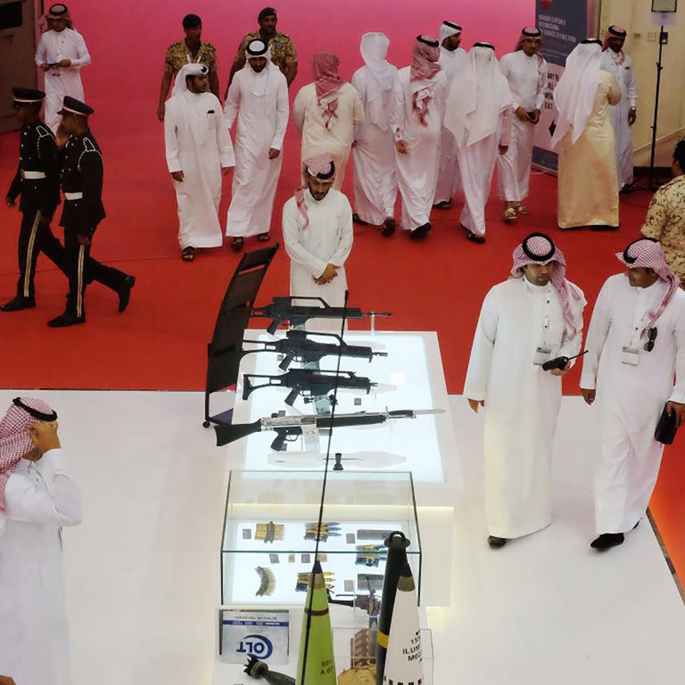 تظهر في الصورة مجموعة من الرجال العرب يرتدون الأزياء التقليدية، يتجولون في مكان يظهر فيه عرض للأسلحة. في وسط الصورة، توجد حاوية زجاجية تحتوي على عدة أنواع من الأسلحة، بما في ذلك البنادق. في الخلفية، يظهر بعض الأفراد المناسبين، مما يوحي بأن الحدث قد يكون معروضًا أو معرضًا للأسلحة أو الأمن. الأجواء تبدو رسمية وجادة، مع وجود السجادة الحمراء في الأرضية.