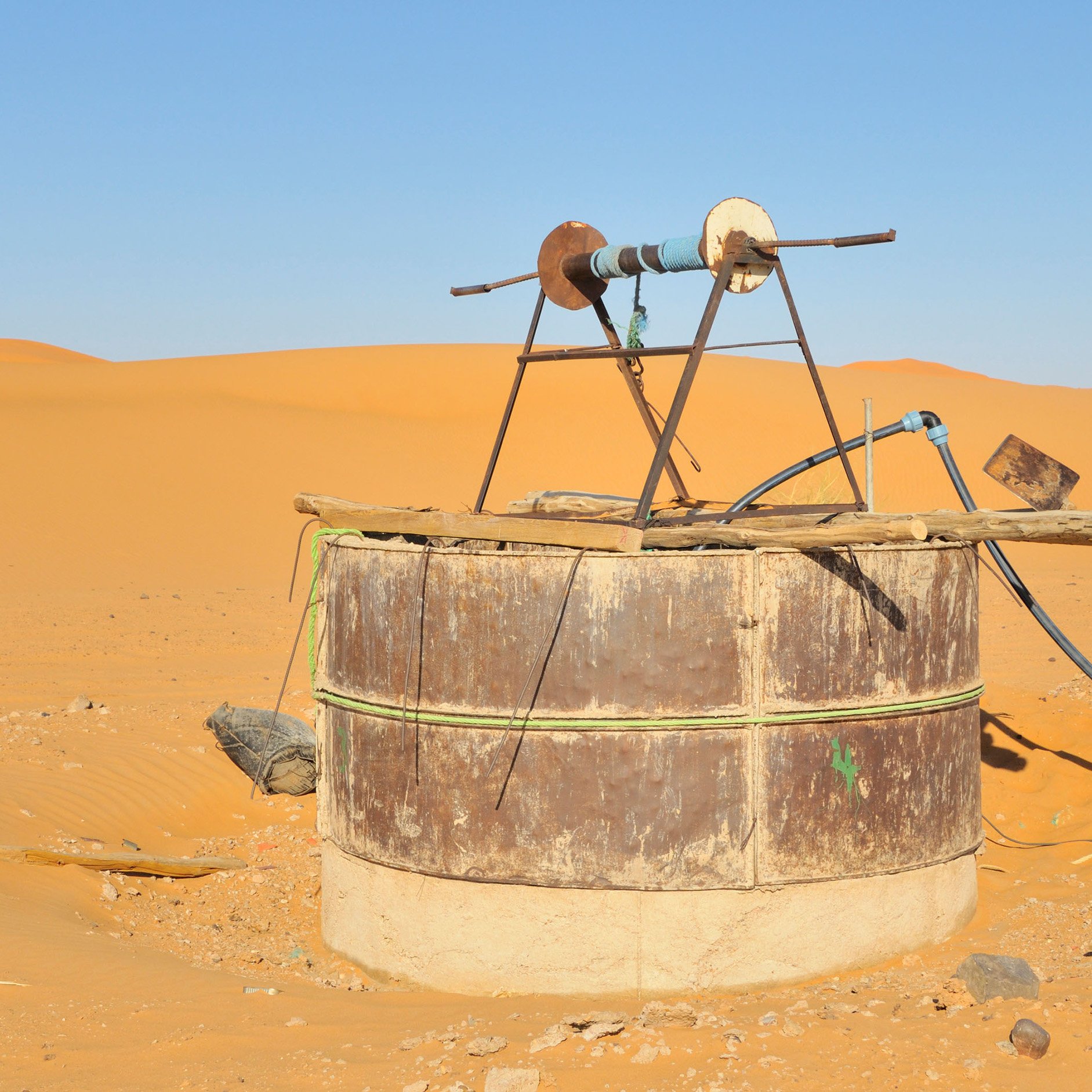 The image shows an old water well situated in a vast desert landscape. The well is made from a large, cylindrical structure with a rusty metal exterior and a concrete base. At the top, there is a simple wooden frame holding a pulley and a couple of wheels, likely used for drawing water. Surrounding the well are sand dunes, displaying a warm golden color under a clear blue sky, emphasizing the arid environment.