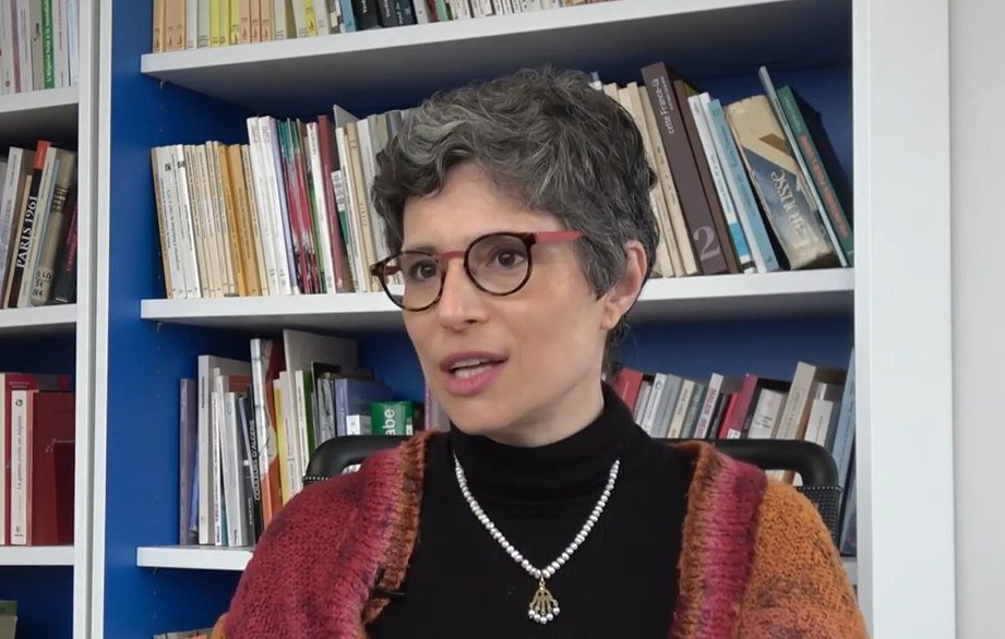 L'image montre une femme avec des cheveux courts et grisonnants, portant des lunettes et un pull noir. Elle est assise devant une bibliothèque remplie de livres, avec un mur bleu derrière elle. Elle a un collier en perles et semble être en train de parler ou de répondre à une question, affichant une expression réfléchie.