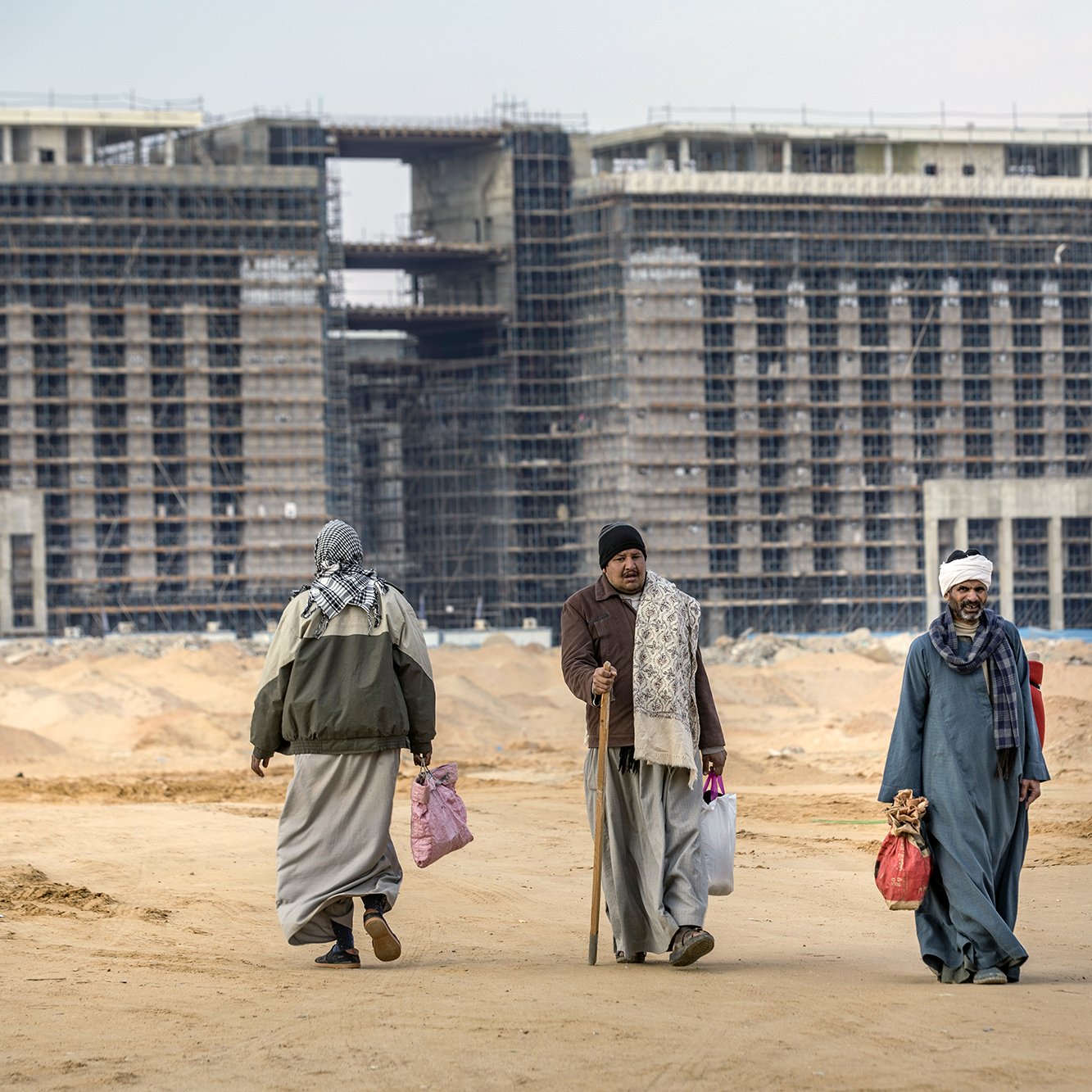 The image depicts a construction site with large, partially completed buildings in the background. In the foreground, four individuals are seen walking on a sandy terrain. They are dressed in traditional attire, including long robes and head coverings. Some are carrying bags, and one person is using a cane. The overall scene reflects a blend of activity and the unfinished nature of the construction, suggesting a developing urban area.