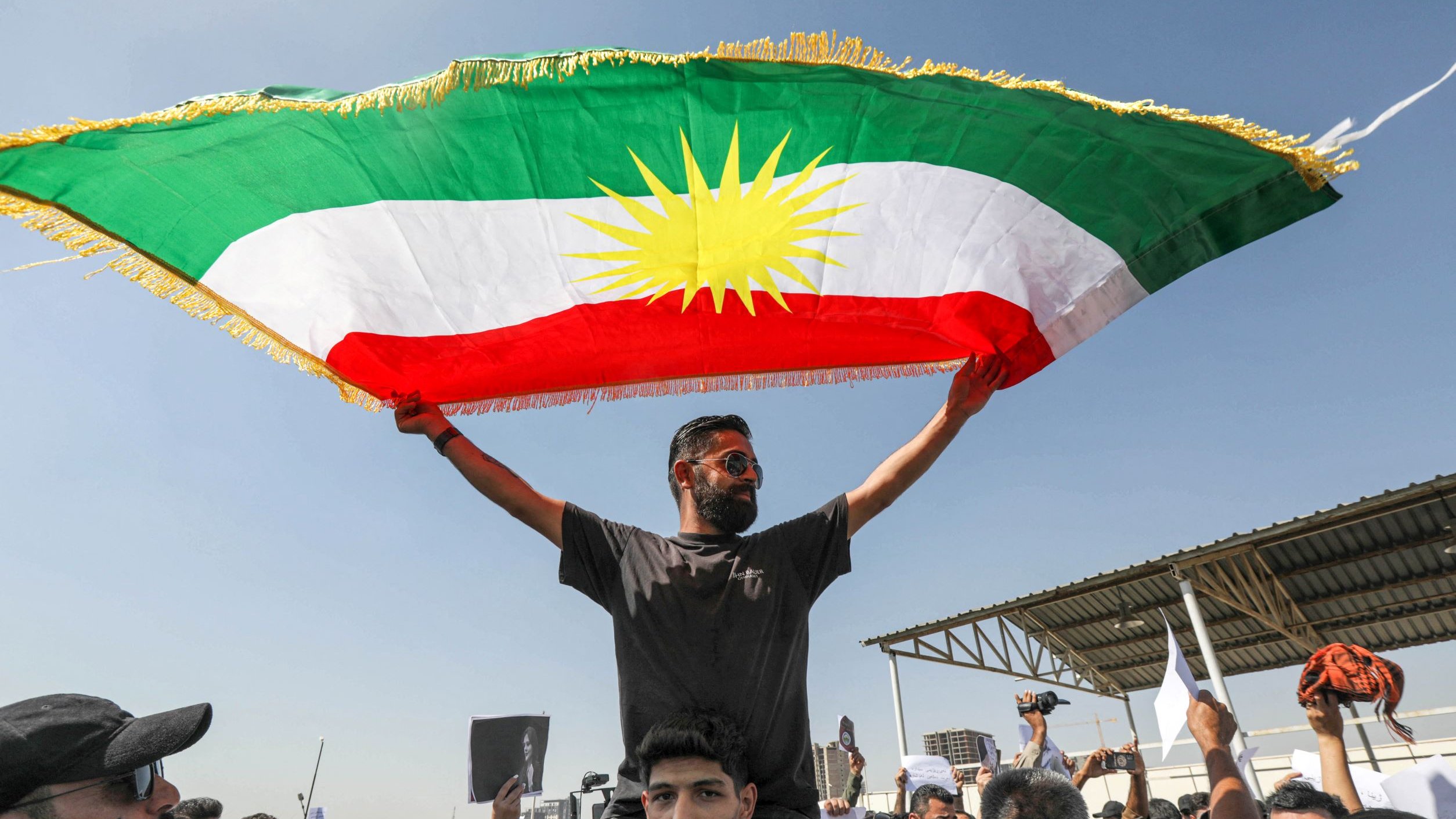 L'immagine mostra un gruppo di persone riunite in una manifestazione. Un uomo, visibilmente emozionato, è in piedi sulla spalla di un altro, mentre solleva con orgoglio una grande bandiera curda, caratterizzata da un fondo verde, rosso e giallo, con un sole stilizzato al centro. Il contesto appare festoso e di protesta, con altre persone intorno che mostrano cartelli e manifestano il proprio sostegno. Sullo sfondo si può notare un'area aperta con una struttura.