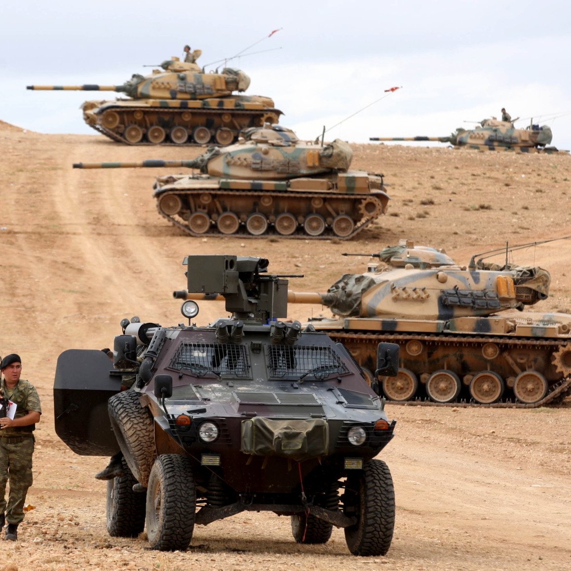 L'image montre un paysage militaire avec plusieurs chars d'assaut en arrière-plan, alignés sur une colline. Au premier plan, un véhicule blindé est garé sur un chemin poussiéreux, tandis qu'un soldat semble faire une inspection ou prendre des notes à côté du véhicule. Le terrain est désertique, et le ciel est partiellement nuageux, créant une atmosphère tendue et militaire. Les chars sont camouflés avec des motifs de peinture qui se fondent dans le paysage environnant.