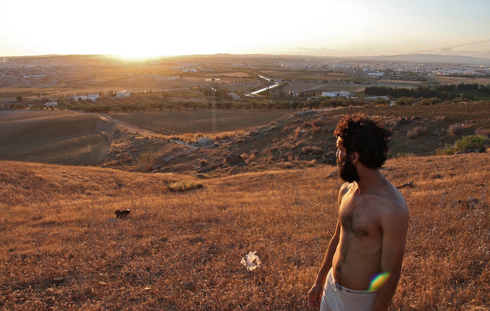L'image montre un homme debout sur une colline, regardant vers un paysage vallonné au coucher du soleil. Il a les cheveux et la barbe bruns, et porte un t-shirt. Le terrain autour de lui est sec et couvert d'herbes hautes. En arrière-plan, on peut voir un petit village et des champs qui s'étendent à perte de vue, créant un panorama paisible et naturel. La lumière du soleil met en valeur les couleurs dorées de la scène.
