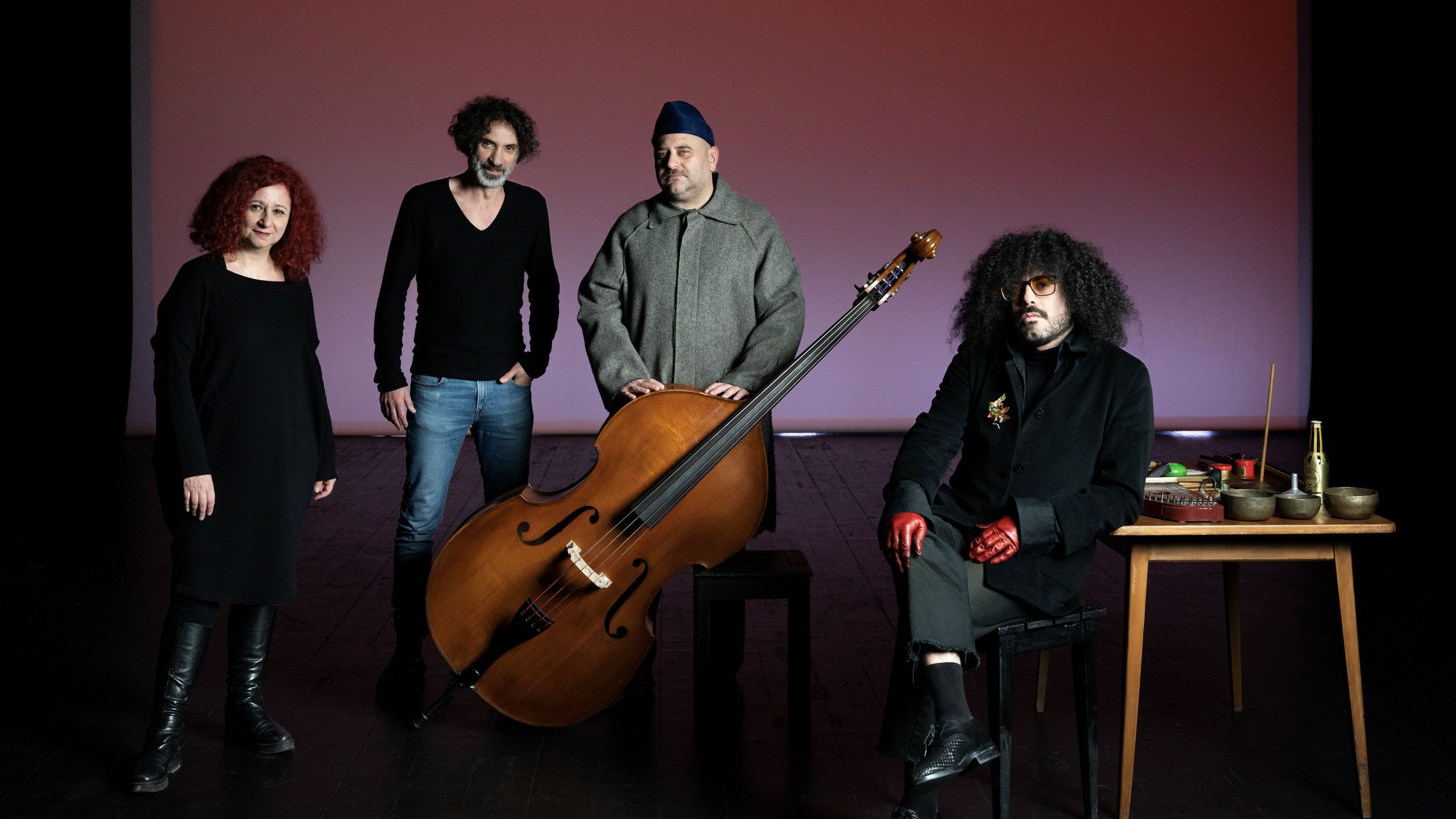 L'image présente un groupe de quatre personnes posant ensemble sur scène. Au centre, il y a un grand instrument de musique, probablement un instrument à cordes, qui attire l'attention. À gauche, une femme avec des cheveux roux bouclés se tient avec une expression sérieuse. À côté, un homme avec des cheveux frisés et un pull noir semble également concentré. Au centre, près de l'instrument, un homme porte une tenue grise et un couvre-chef. Enfin, à droite, un autre homme avec des lunettes et des gants rouges est assis à une table avec divers objets, ayant l'air intrigant. L'éclairage de la scène crée une ambiance artistique et captivante.