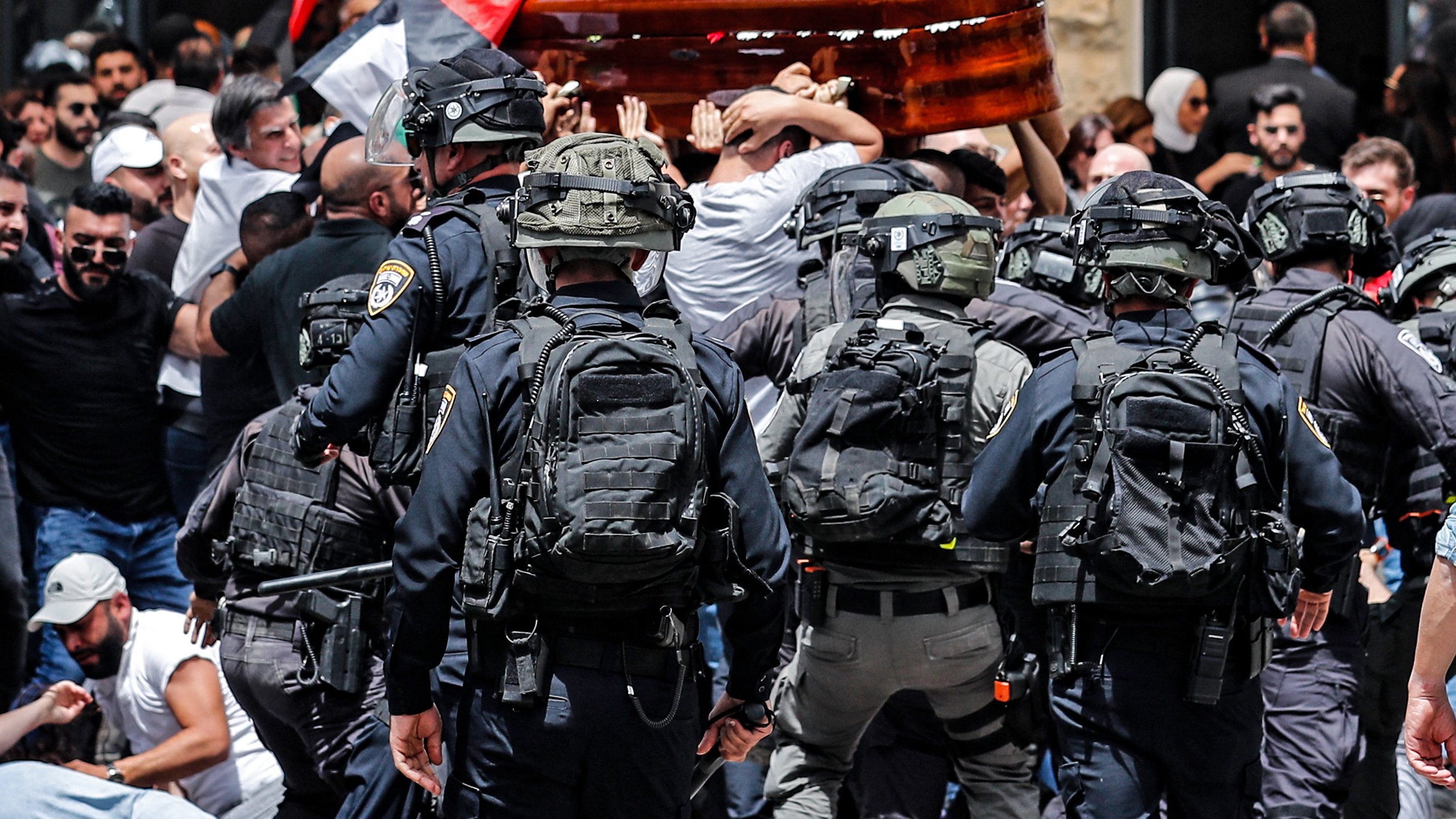The image depicts a scene of tension and conflict. In the foreground, a group of police officers in tactical gear is facing a larger crowd of people who are mournfully carrying a wooden coffin. The crowd appears to be protesting or commemorating a significant event, with flags visible, including Palestinian flags. The atmosphere is charged, suggesting a clash between the police and the public. Emotions are high as both sides engage in this moment.