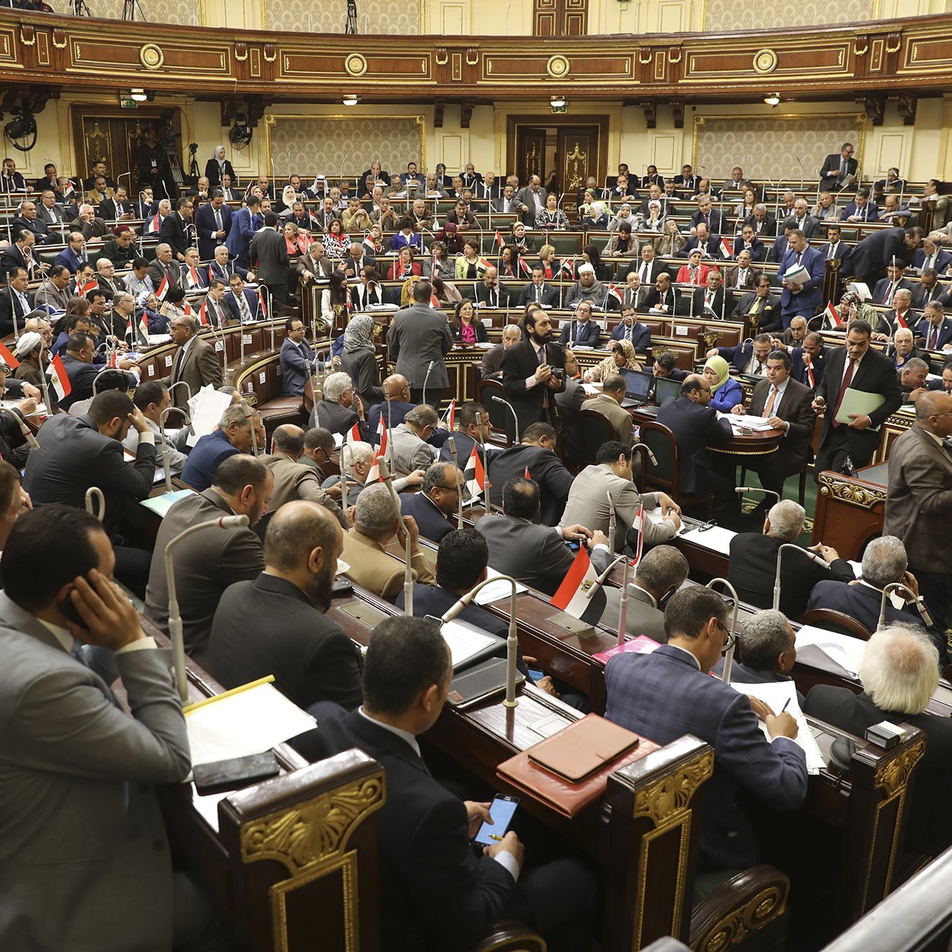 L'image montre une assemblée ou un parlement en pleine session. De nombreux représentants sont assis dans des rangées, certains discutent tandis que d'autres prennent des notes ou consultent des documents. On peut voir des drapeaux égyptiens sur certaines tables, ce qui suggère que cette image provient de l'Assemblée nationale d'Égypte. L'environnement est formel, avec une riche décoration et une disposition typique d'une salle de réunion politique.