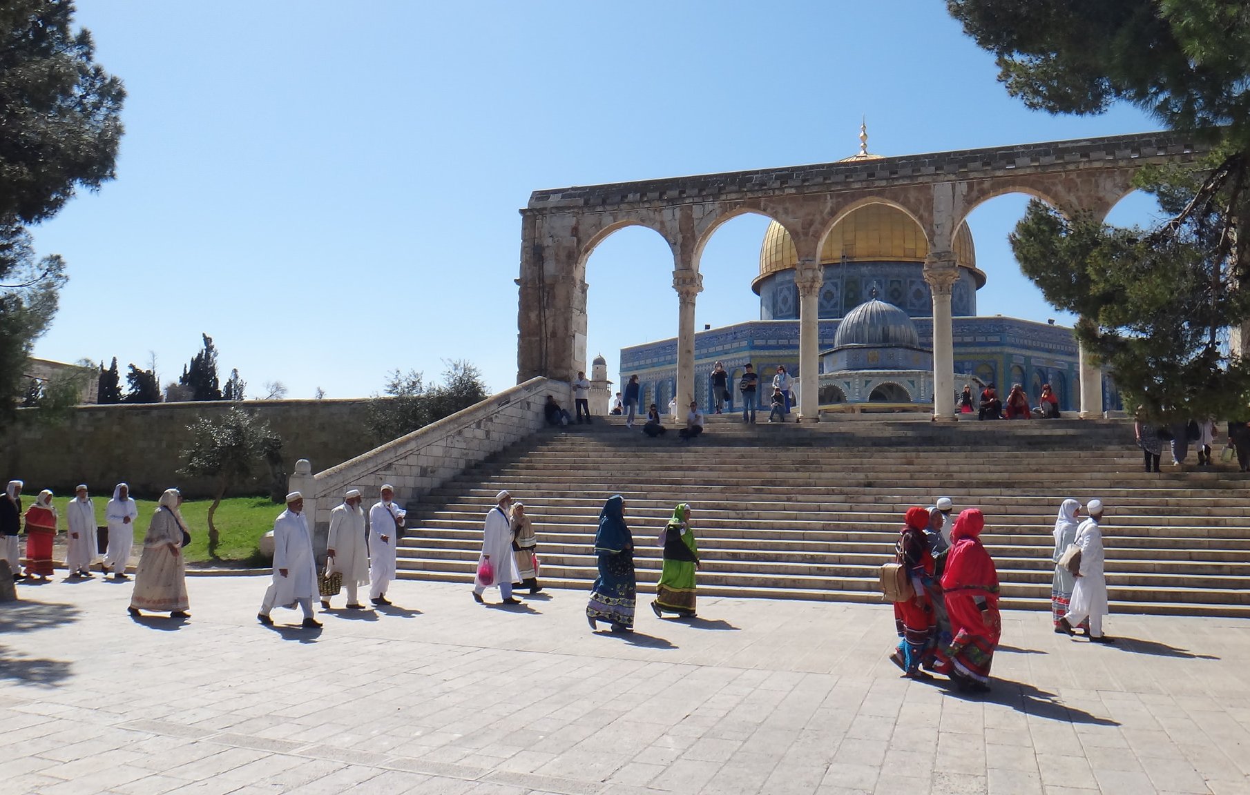 L'image montre un paysage architectural impressionnant avec des arches majestueuses en premier plan. On peut voir un groupe de personnes marchant sur une large marche, certaines portant des vêtements traditionnels colorés. En arrière-plan, la célèbre mosquée avec son dôme doré est visible, entourée d'un ciel clair et d'arbres verdoyants. L'atmosphère semble paisible et spirituelle, reflet d'un lieu de rassemblement important.