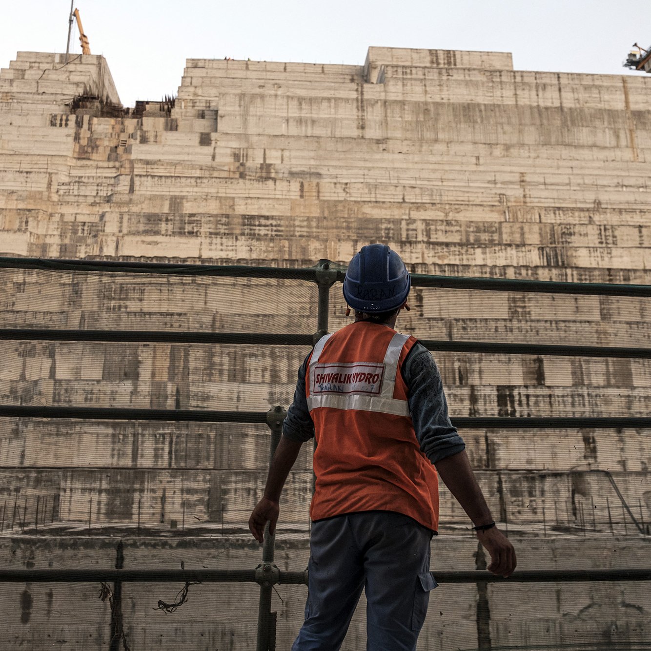 L'image montre un ouvrier sur un chantier de construction, portant un gilet de sécurité orange et un casque. Il se tient devant une grande structure en béton, apparemment inachevée, qui est composée de plusieurs niveaux superposés. En arrière-plan, on aperçoit une grue, ce qui indique que le site est actif. L'atmosphère semble à la fois industrielle et impressionnante, mettant en avant l'ampleur du projet de construction.