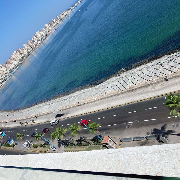 La imagen muestra una vista aérea de una costa bien iluminada. Se puede ver una carretera que corre paralela a la playa, con varios vehículos transitando. A lo largo de la playa hay palmeras, y el agua del mar es de un color azul claro. Al fondo, se pueden observar edificios que forman parte de un área urbana, y el cielo está despejado, lo que sugiere un día soleado.