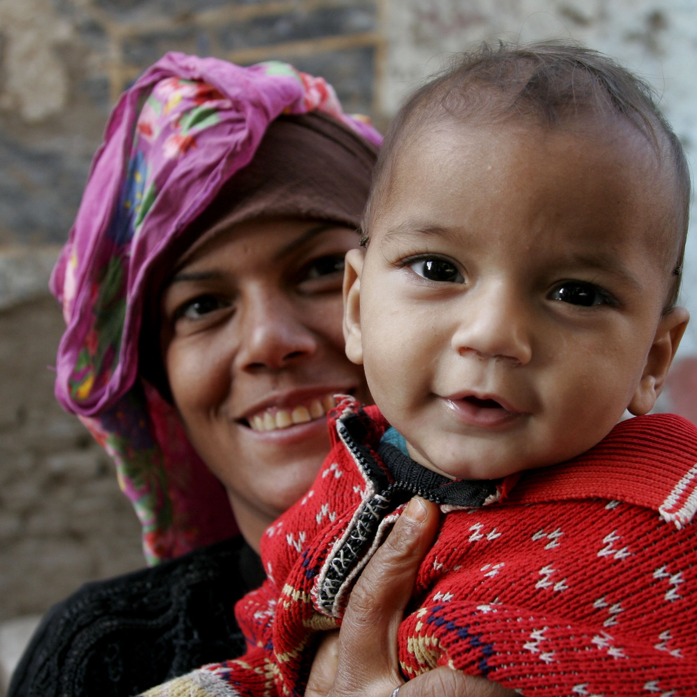 The image features a smiling woman holding a small child. The woman is wearing a colorful headscarf, and her expression is warm and cheerful. The child, who is dressed in a bright red sweater with a patterned design, looks happy and is facing the camera, showing a playful smile. The background appears to be a textured wall, suggesting an outdoor or rustic setting. The overall mood of the image is joyful and intimate, capturing a moment of connection between the woman and the child.
