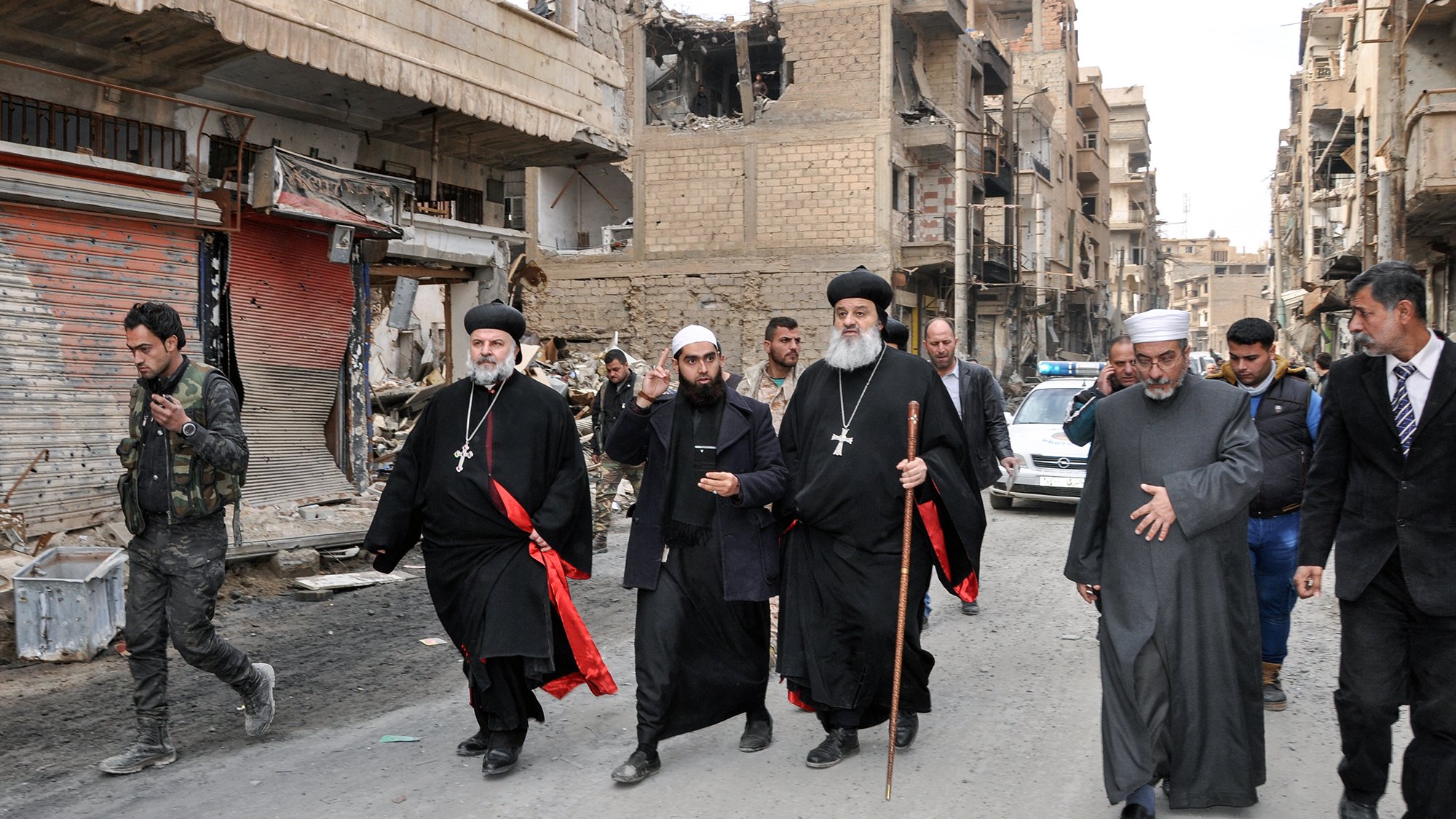 The image shows a group of religious figures walking through a war-torn street. They are dressed in traditional clergy attire, including black robes and crosses, and some are holding staffs. The surroundings are heavily damaged, with buildings showing signs of destruction and debris on the street. In the background, there are a few people and vehicles, indicating a somber atmosphere in a once-bustling area. The overall scene reflects the impact of conflict on the community and highlights the presence of religious leadership amidst the destruction.