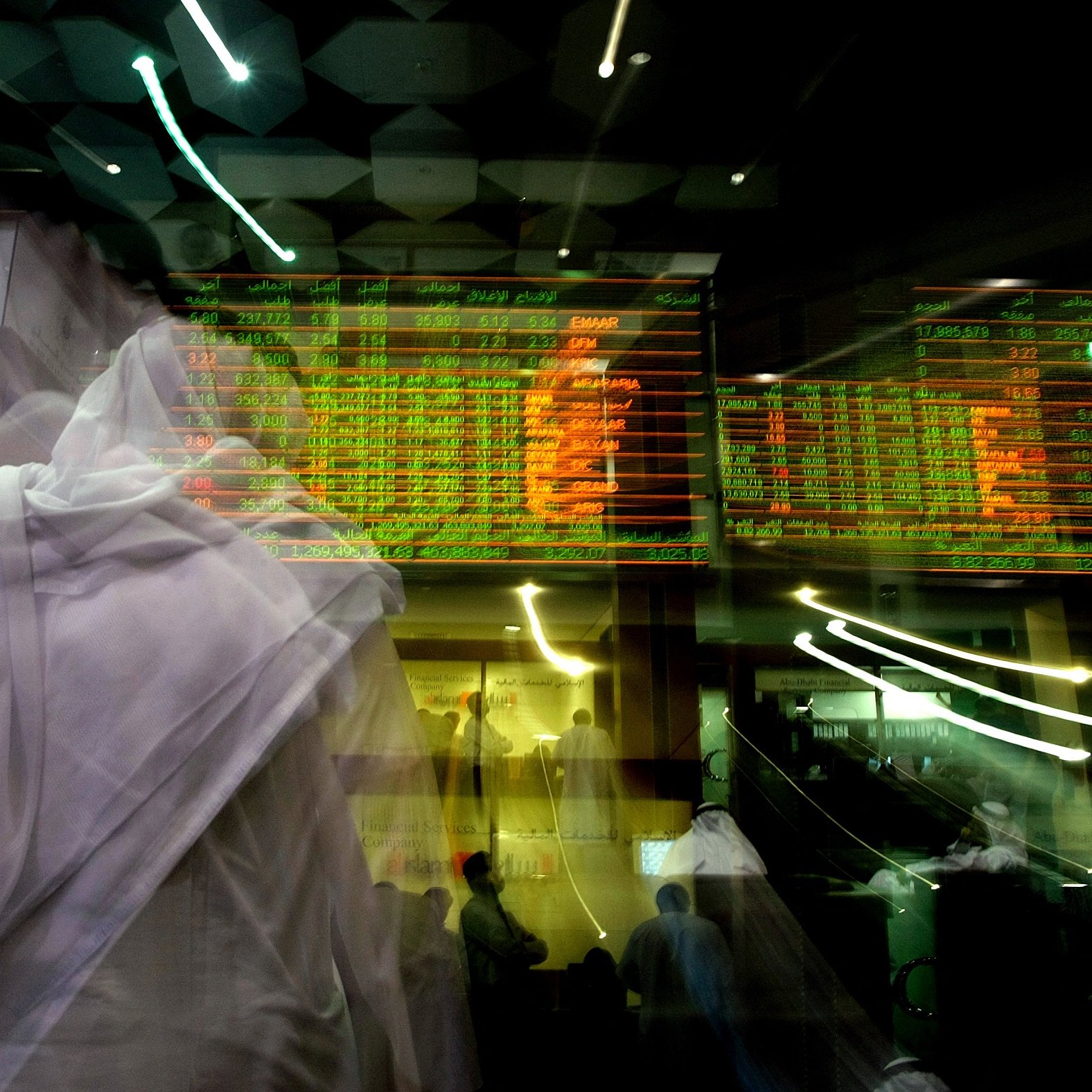 تظهر الصورة مشهدًا من داخل سوق الأسهم، حيث يُرى عدد من الأشخاص يرتدون الملابس التقليدية. الخلفية تحتوي على شاشات تعرض أسعار الأسهم والأرقام، والألوان الساطعة مثل الأخضر والأحمر تشير إلى تغيرات أسعار السوق. تتسم الصورة بالضبابية والحركة، مما يعكس النشاط والحيوية الموجودة في هذا المكان.