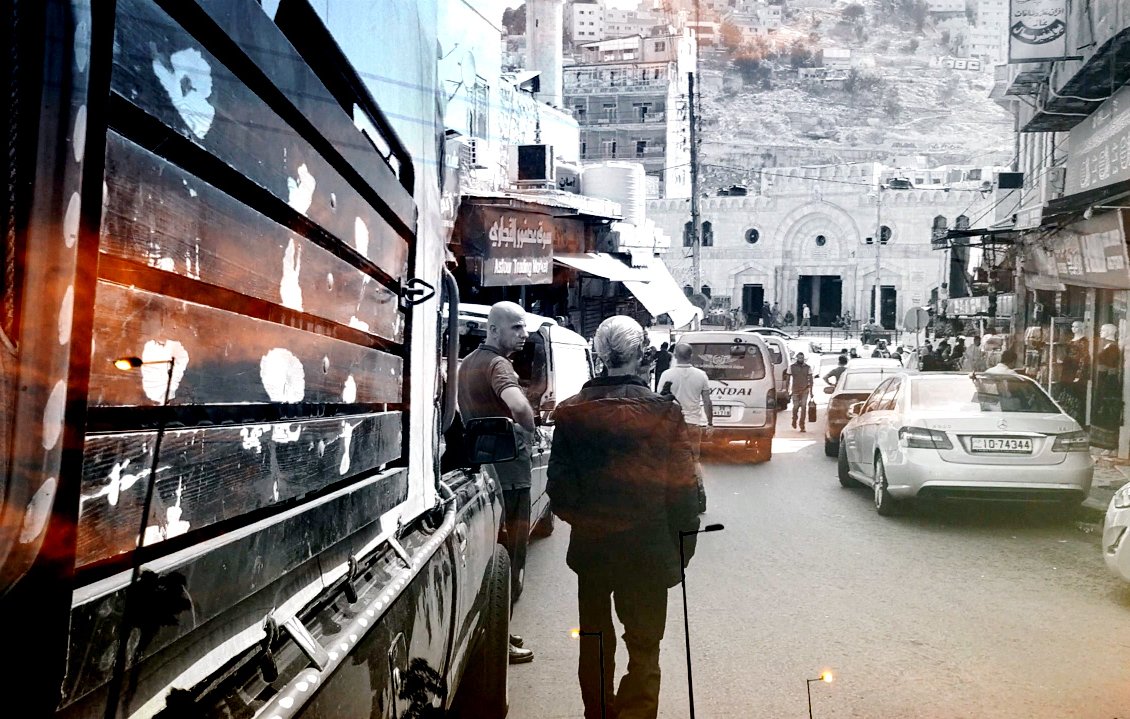 L'image montre une rue animée avec des véhicules garés et des passants. On aperçoit un homme âgé marchant avec une canne, tandis que d'autres personnes se déplacent. À l'arrière-plan, des bâtiments et des collines sont visibles, suggérant un environnement urbain. La photo semble avoir un effet de noir et blanc, avec des touches de couleur chaude, créant une ambiance nostalgique.