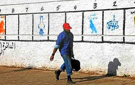 L'image montre un homme marchant le long d'un mur peint en blanc. Sur le mur, il y a une série de numéros de 1 à 8 avec des illustrations ou des symboles correspondant à chaque numéro. L'homme porte une casquette rouge et un vêtement bleu, tenant une mallette. À gauche du mur, une inscription en arabe est visible. L'environnement semble urbain et ensoleillé.
