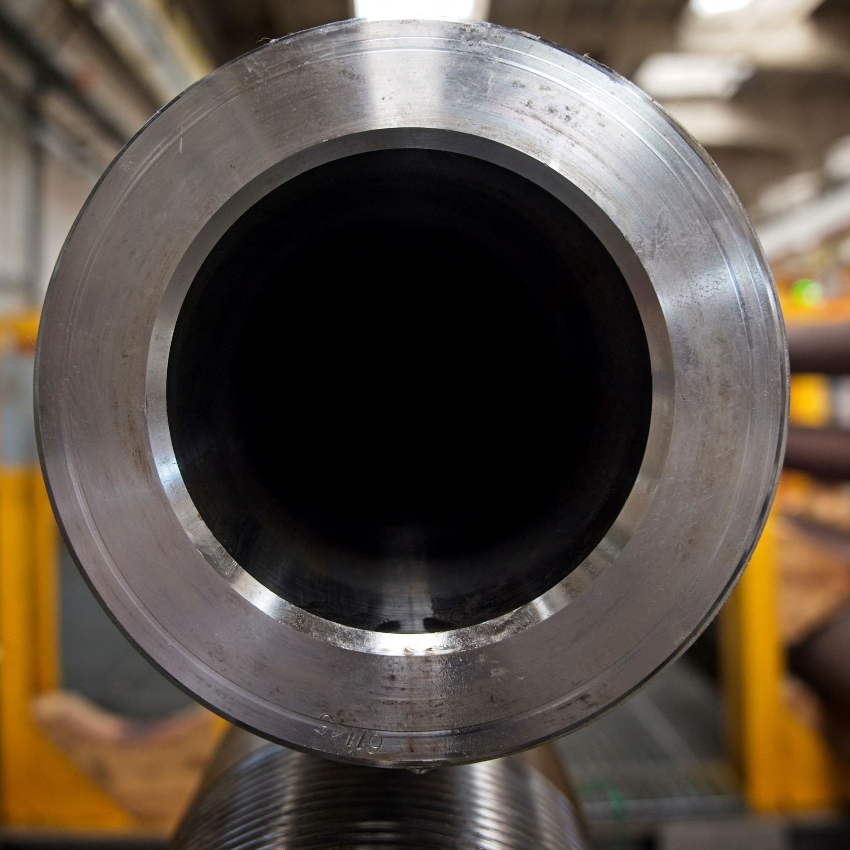 تظهر في الصورة أنبوب معدني كبير، حيث يتمركز التركيز على فتحة الأنبوب من الداخل. الجوانب الخارجية للأنبوب تظهر لامعة، بينما الداخل مظلم تمامًا. خلف الأنبوب، يمكن رؤية بعض العناصر الصناعية التي تشير إلى وجود مصنع أو ورشة عمل. الإضاءة في المكان تبدو طبيعية، مما يضفي على الصورة جوًا صناعيًا.