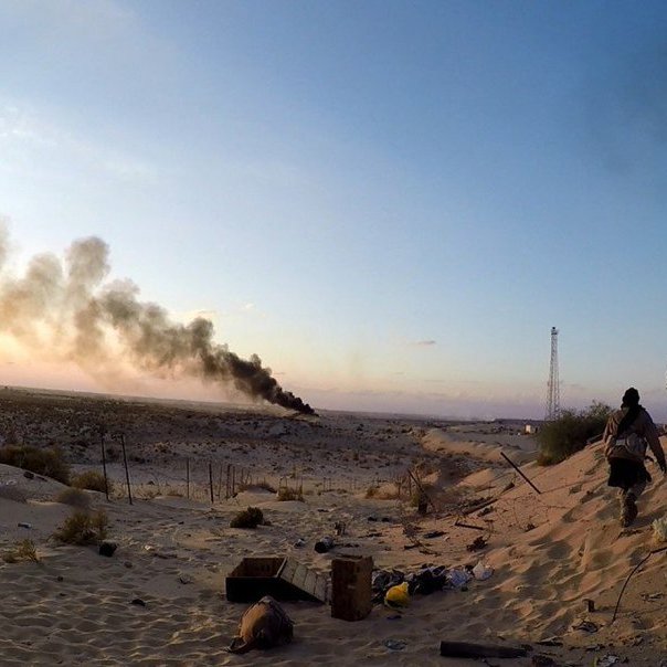 L'image montre un paysage désertique avec une ambiance dramatique. À l'arrière-plan, on peut voir des colonnes de fumée noire s'élevant dans le ciel, suggérant qu'il y a un incendie ou une explosion. Au premier plan, des silhouettes de personnes évoluent dans le sable, probablement en train d'explorer ou de surveiller la zone. La lumière dorée du coucher de soleil crée un contraste frappant avec l'obscurité de la fumée, ajoutant à l'atmosphère générale de tension et de désolation. Le décor semble abandonné, avec quelques débris éparpillés sur le sol.