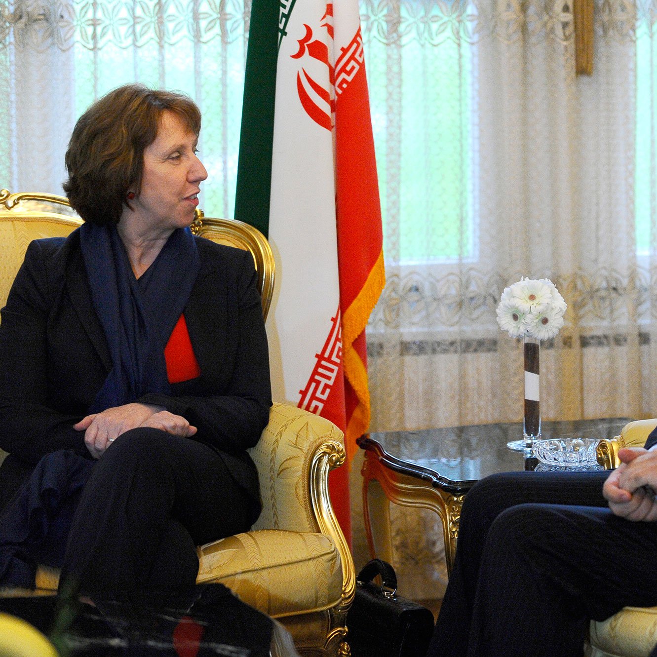 L'image montre une rencontre entre deux personnes assises sur des fauteuils dans un décor formel. À gauche, une femme avec des cheveux bruns, portant un blazer noir et un haut rouge, semble engagée dans la conversation. À droite, un homme souriant, vêtu d'un costume sombre, répond à la femme. En arrière-plan, on peut voir un drapeau iranien, ce qui suggère que la rencontre a lieu en Iran. La lumière naturelle entre par les fenêtres, créant une atmosphère accueillante.
