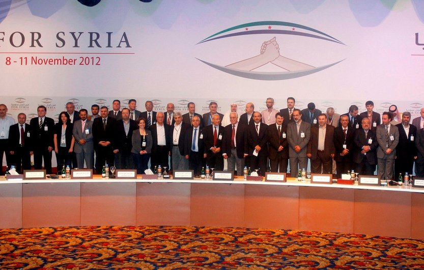 L'image montre un groupe de personnes réunies lors d'un événement formel, probablement une conférence. Elles sont alignées en plusieurs rangées, toutes en costume, et semblent participer à un rassemblement lié à la Syrie, comme indiqué par le texte en arrière-plan. Le décor est coloré, avec des drapeaux et des motifs qui évoquent l'événement. Les participants adoptent des expressions sérieuses, ce qui suggère l'importance de l'occasion.