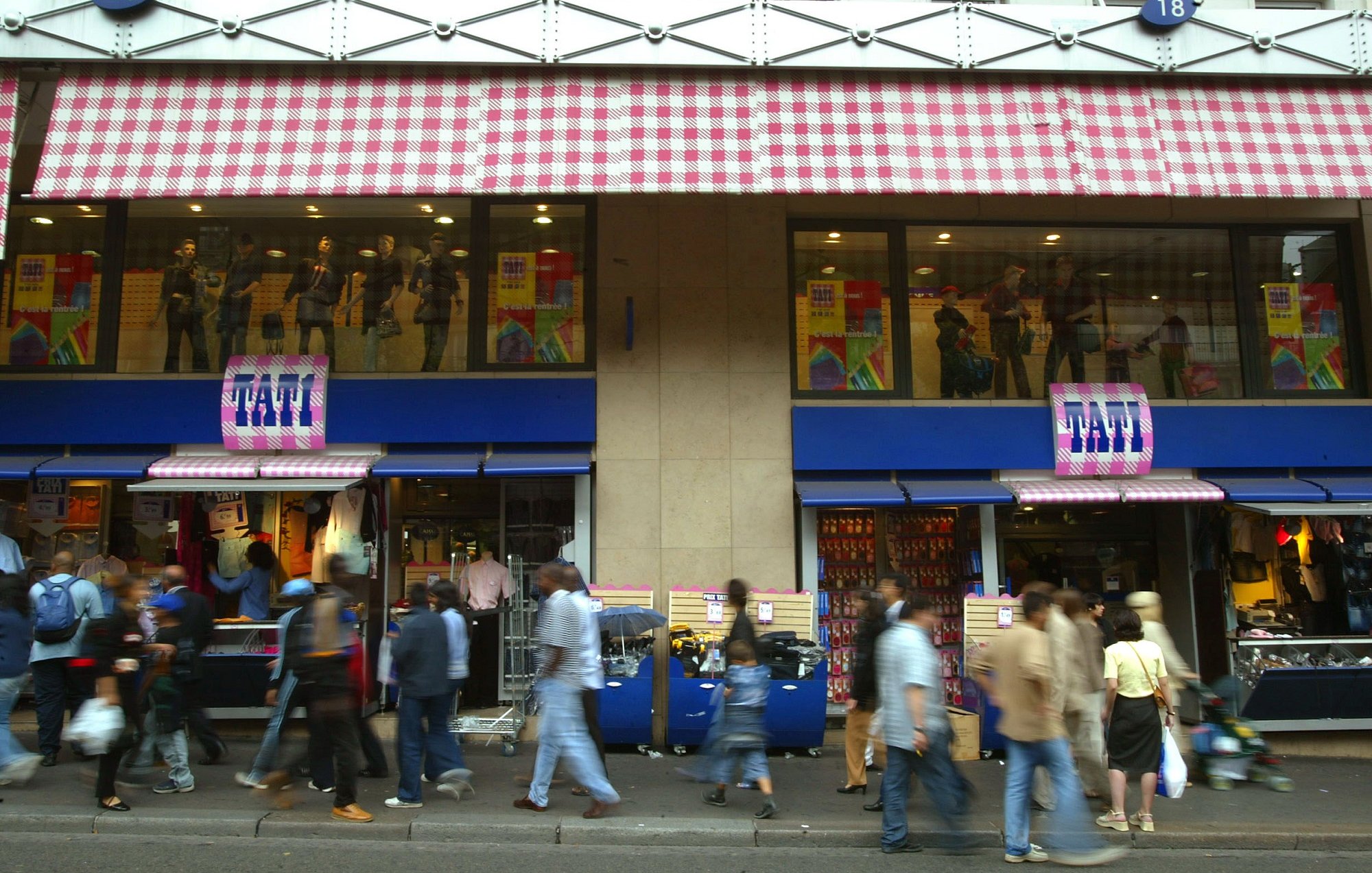 L'image montre une rue animée avec des piétons marchant devant un magasin. Le magasin est identifié par un enseigne "FATI" et présente des vitrines avec des mannequins habillés. Les façades supérieures du magasin ont des stores à carreaux. On peut voir une multitude de personnes se déplacer sur le trottoir, créant une atmosphère dynamique et vivante.