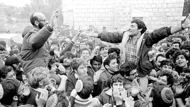 L'image montre une foule de personnes rassemblées, avec des visages enthousiastes et des expressions de joie. Deux hommes se distinguent, l'un portant des lunettes, debout sur les épaules de l'autre, qui lève les bras en signe de célébration. Les membres de la foule semblent excités et engagés, et l'environnement est peut-être urbain ou rural, entouré de bâtiments en pierre. L'atmosphère générale évoque un moment de rassemblement collectif et de camaraderie.