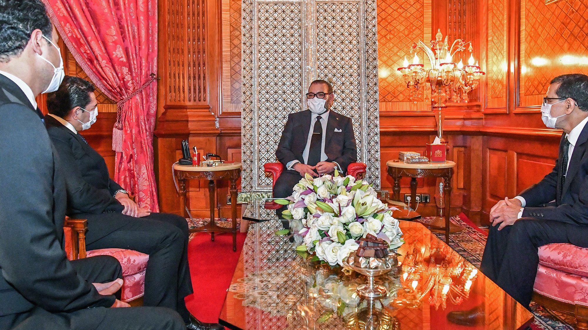 L'image montre un intérieur élégant avec un décor riche. Au centre, une table en verre repose sur un tapis rouge. On voit un homme assis, probablement un leader ou une personnalité importante, entouré de trois autres personnes, toutes en tenue formelle. Ils portent des masques, suggérant que la rencontre se déroule dans un contexte de précautions sanitaires. Le fond présente des murs ornés et un espace bien éclairé, avec des éléments décoratifs comme des fauteuils et des fleurs sur la table.