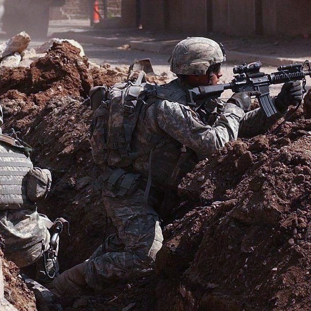 L'image montre des soldats en engagement militaire, se cachant derrière des tranchées. Ils portent des uniformes militaires et des casques, et sont armés. Le terrain est marqué par des gravats et de la terre, suggérant un environnement de combat. L'atmosphère est tendue, montrant la préparation et la vigilance des soldats face à un danger.