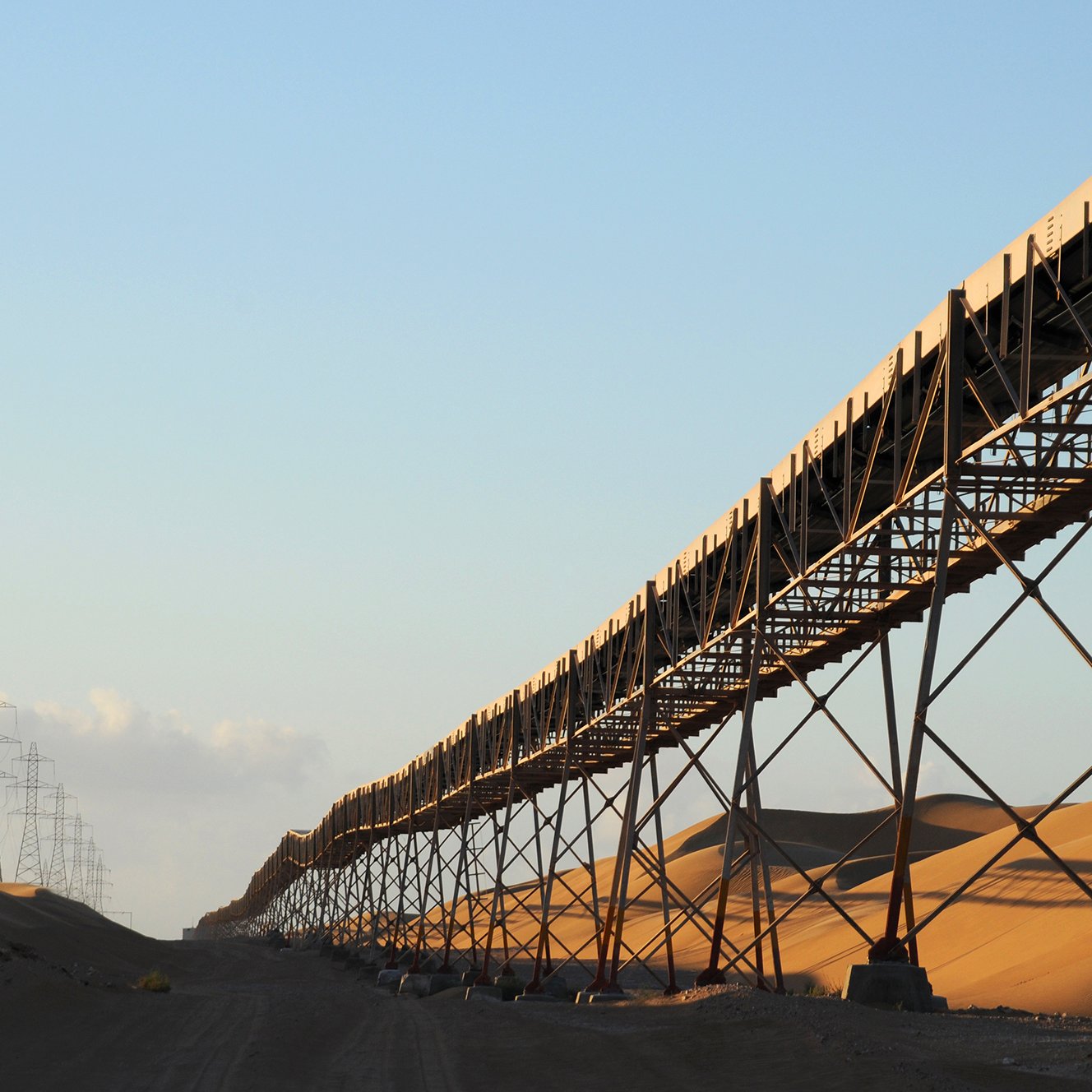 The image depicts a long conveyor belt system stretching across a vast landscape dominated by sand dunes. The conveyor belt is elevated on metal supports, running parallel to the ground. In the background, tall power lines extend into the horizon, hinting at an industrial setting. The warm light of the sun creates a golden hue on the sand, adding to the serene yet striking composition of the scene. The overall atmosphere suggests a remote, arid environment that may be associated with mining or agricultural operations.