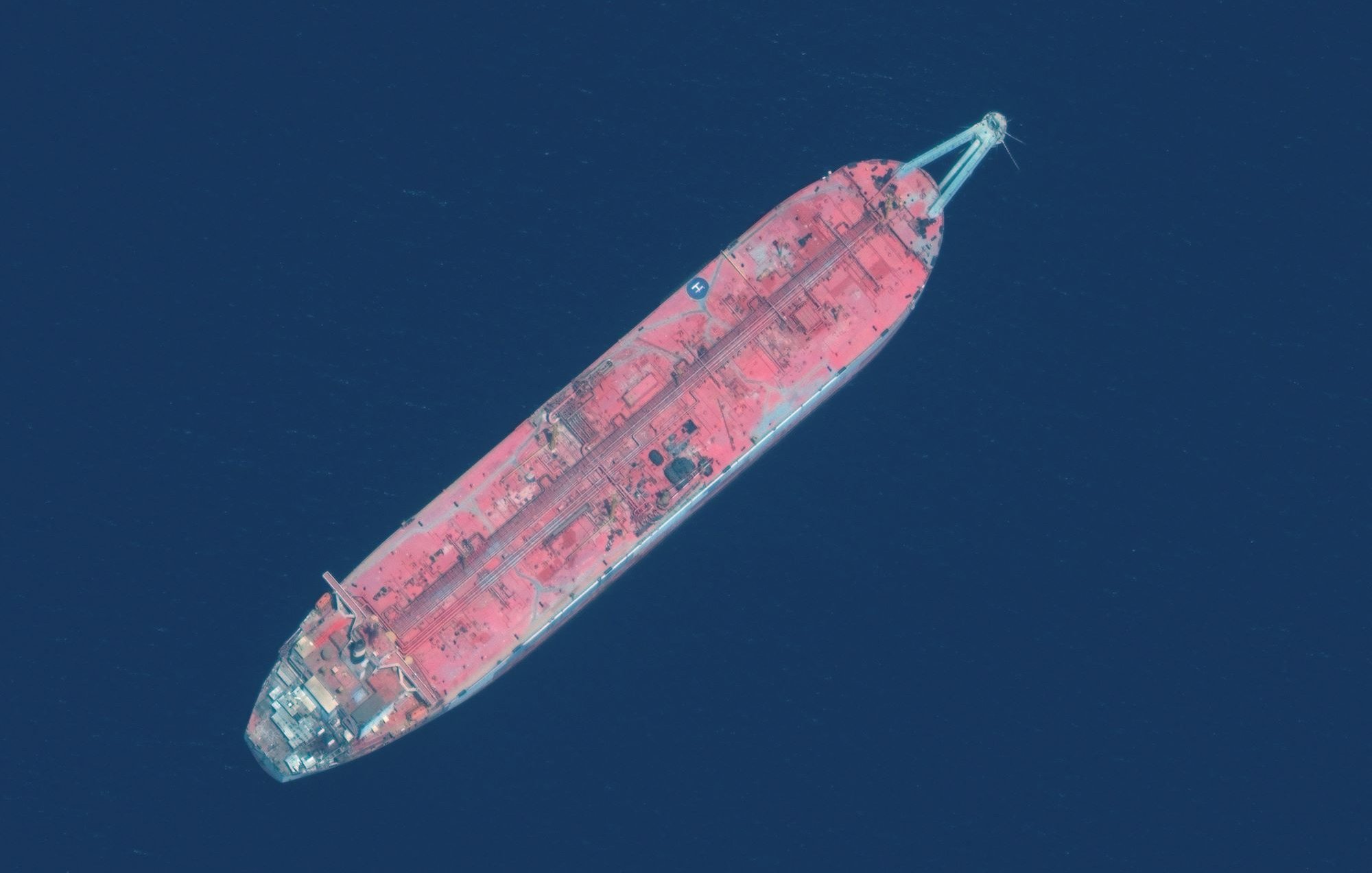 L'image montre un grand navire de transport de pétrole, vu du dessus. Le navire a une coque de couleur rouge, avec des détails visibles sur le pont. Il flotte sur une surface d'eau bleue profonde, ce qui donne un fort contraste entre le navire et son environnement. Les structures sur le pont semblent usées, suggérant un certain âge ou une exposition aux éléments marins.