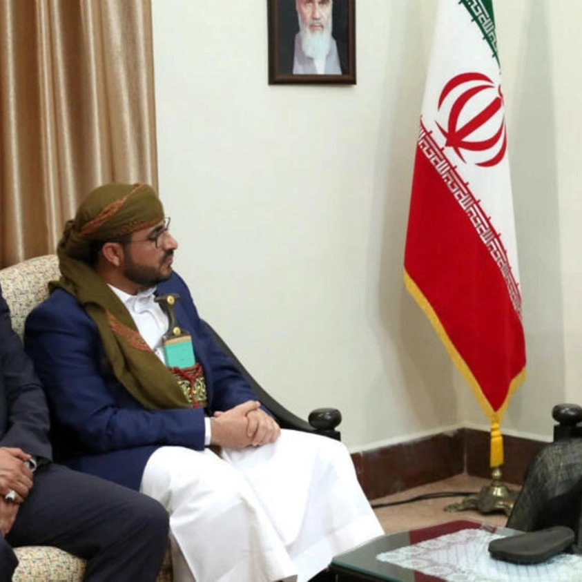 الصورة تُظهر ثلاثة رجال يجلسون في غرفة اجتماعات. في الخلفية، يوجد علم إيران وصورة كبيرة على الحائط. الرجل الموجود على اليمين يرتدي زيًا تقليديًا ويبدو أنه يتحدث مع الرجلين الآخرين، الذين يجلسون أمامه. يُظهر تعبيرهم اهتمامًا وتفاعلًا مع الحديث. الغرفة تبدو رسمية، مع أثاث بسيط وإضاءة دافئة.