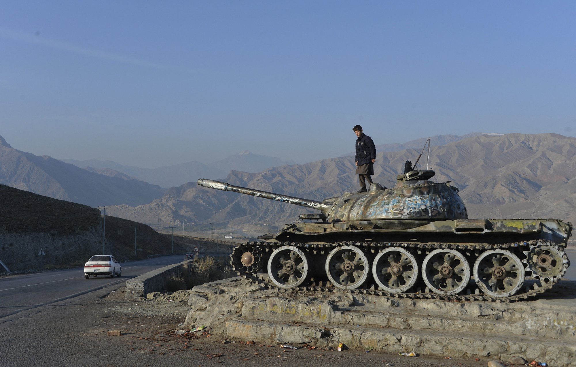 L'image montre un homme debout sur un char de bataille, probablement un ancien char T-55, qui est posé sur une plateforme. Le char présente des signes d'usure et de rouille, et le paysage environnant est montagneux avec un ciel dégagé. On peut voir une route à proximité, ainsi qu'une voiture blanche circulant. L'ambiance évoque un mélange de nostalgie et de désolation, rappelant une époque passée de conflits.