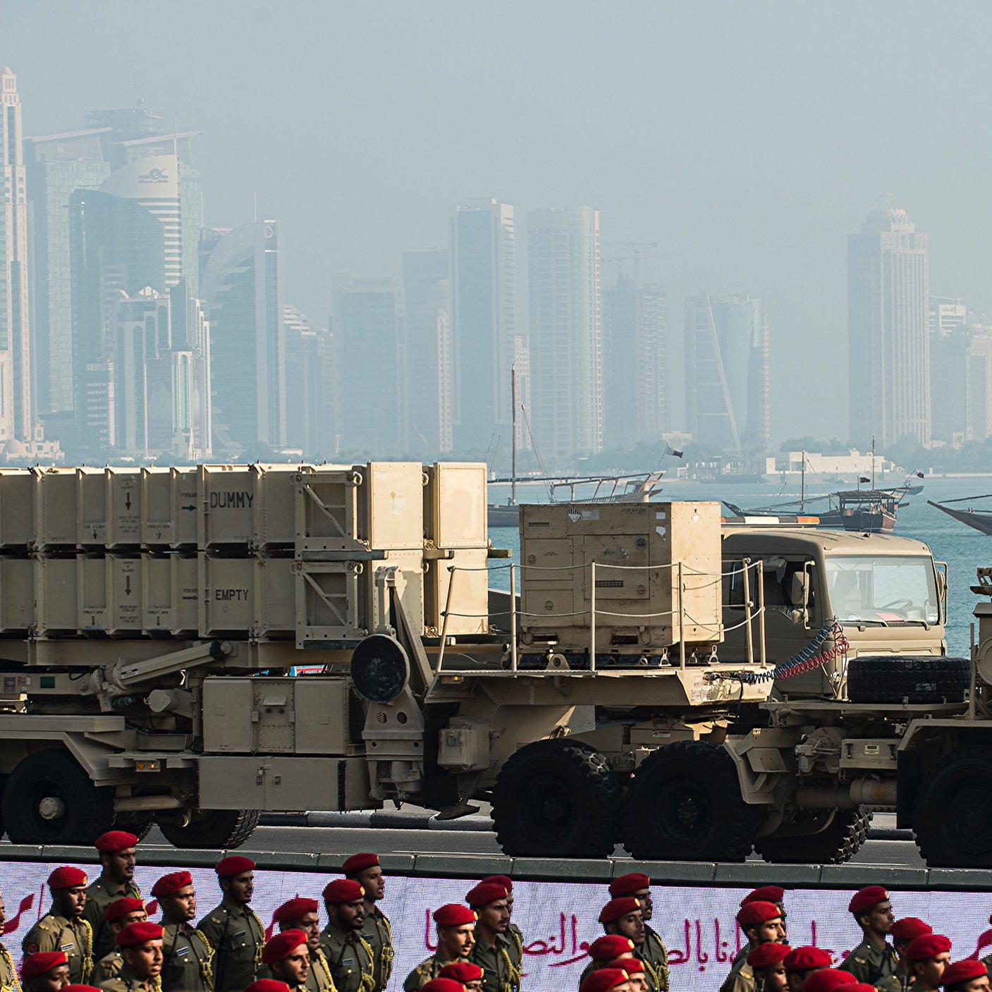 L'image montre des équipements militaires, probablement des systèmes de défense aérienne, positionnés sur des camions. En arrière-plan, on aperçoit une ville moderne avec des gratte-ciels et des bâtiments contemporains, ainsi qu'une étendue d'eau et des bateaux traditionnels. Au premier plan, des soldats en uniforme, peut-être d'une force armée, sont alignés, ajoutant une dimension cérémonielle à la scène. L'atmosphère semble être celle d'un défilé militaire ou d'un événement similaire.